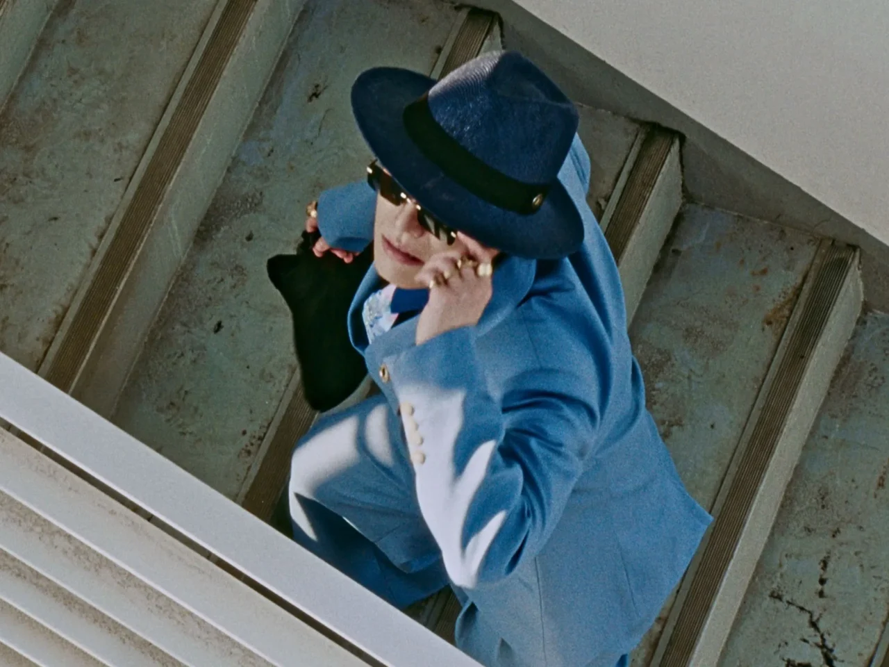 Person in a blue suit and matching hat adjusts sunglasses while standing on concrete stairs.