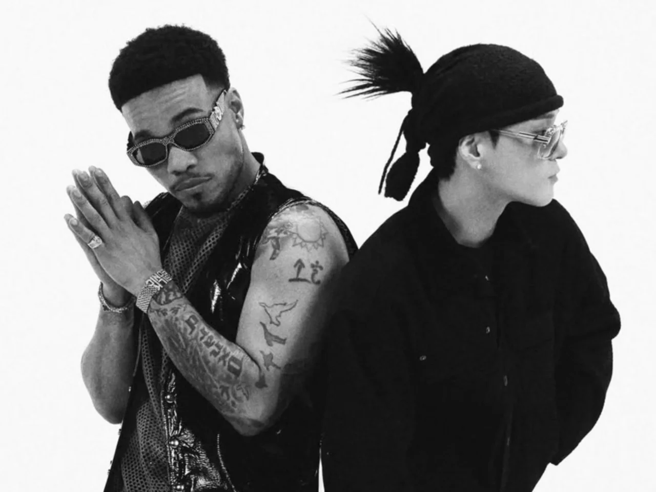 Two people stand back-to-back in a black-and-white photo; left is a tattooed man wearing sunglasses and clasping his hands, right a woman in a hat and sunglasses.