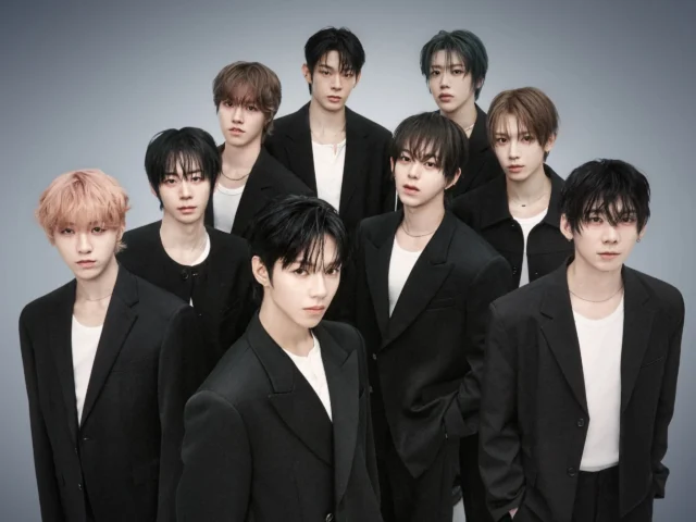 Nine men in black blazers pose in a studio against a pale blue backdrop.