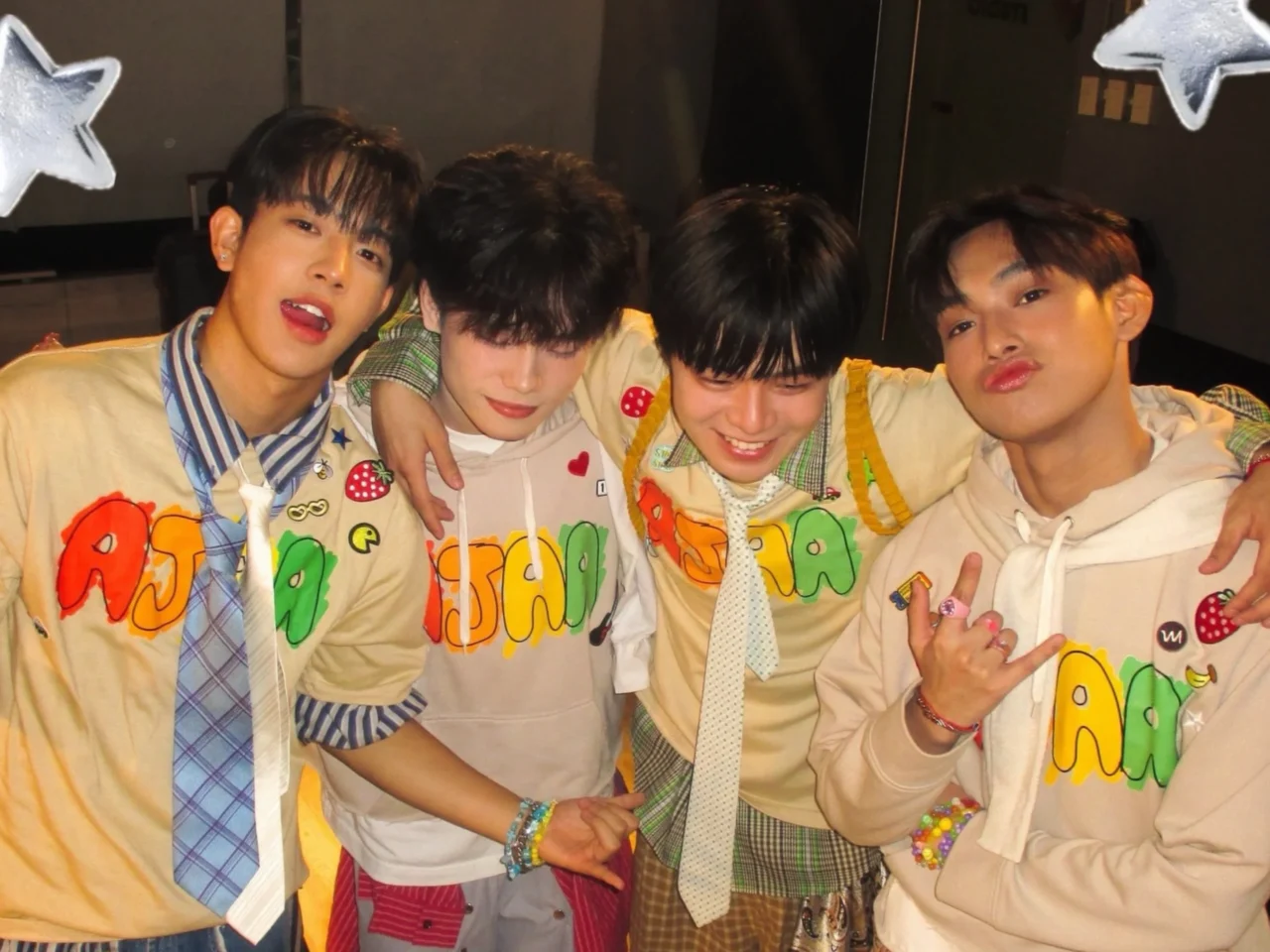 Four young men pose together with arms around each other, smiling at the camera in colorful outfits with graphic letters and patches.