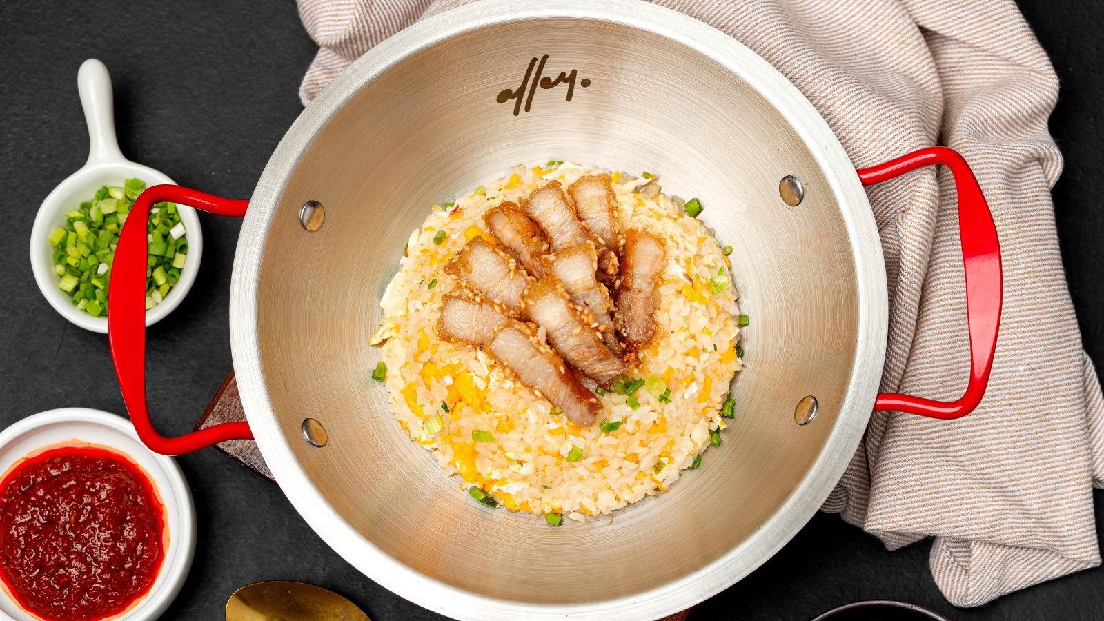 Pan of fried rice with sliced pork on top, green onions, and a red-handled pot, surrounded by condiments and a striped cloth.
