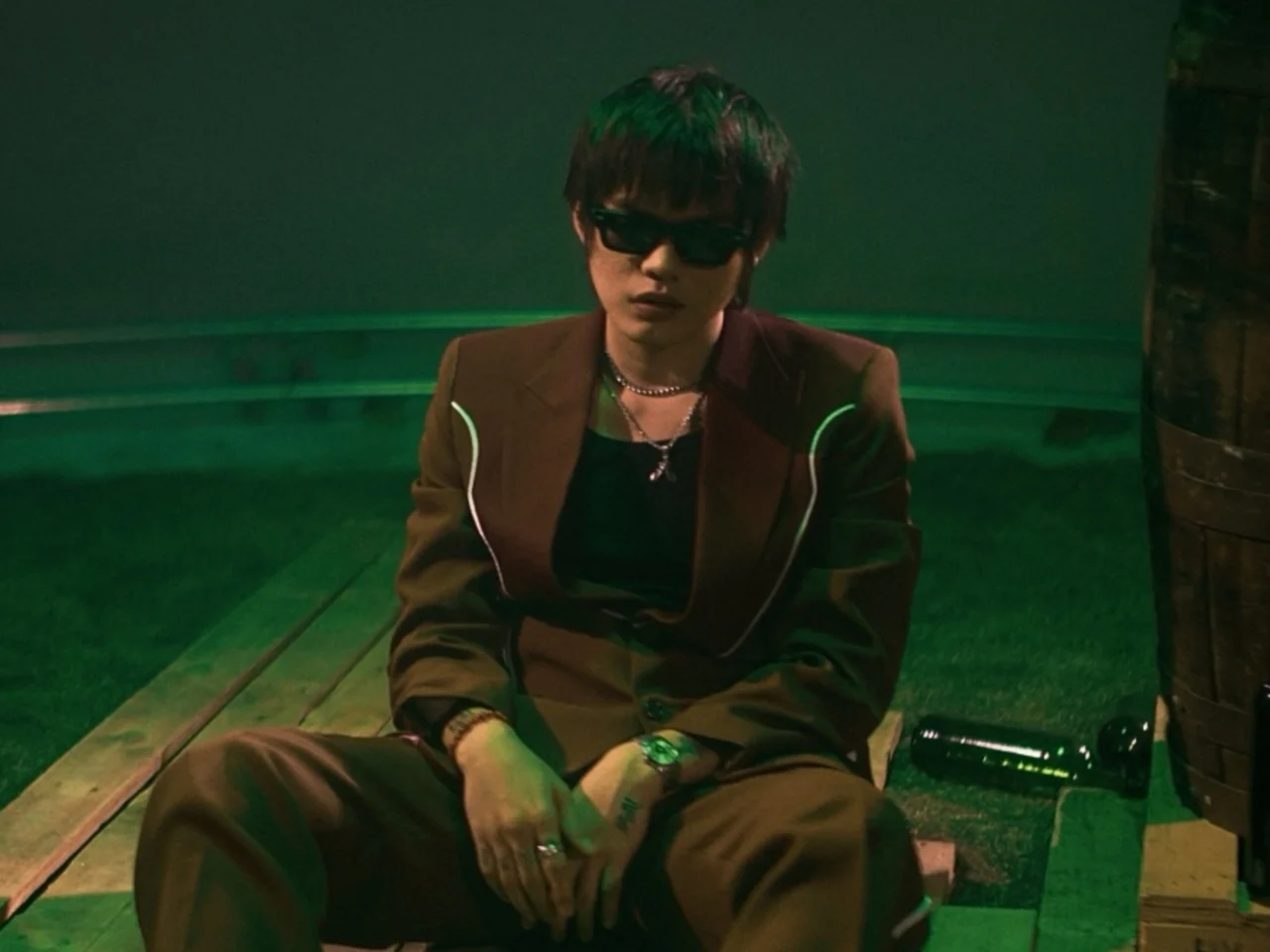 Man in a brown blazer and dark sunglasses sits on wooden steps under green lighting, facing the camera.
