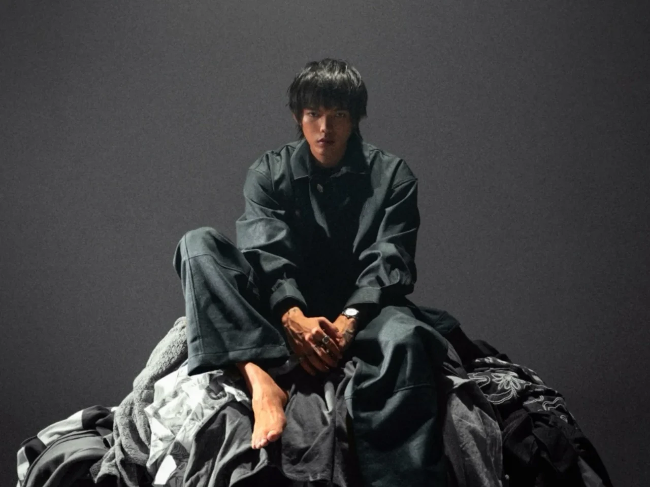 Young man seated in a dark studio wearing a dark denim outfit, looking at the camera.