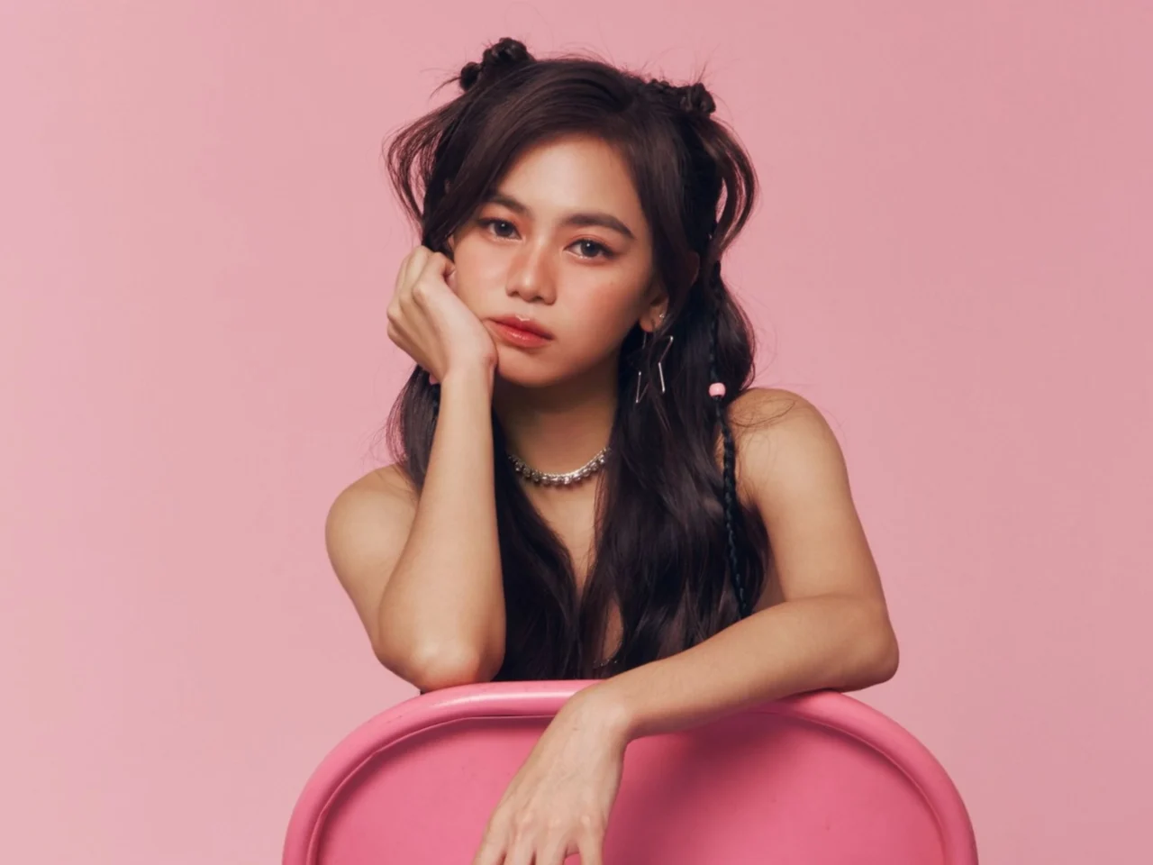 Young woman with long dark hair resting her chin on her hand against a pink backdrop, wearing hoop earrings and a silver necklace.