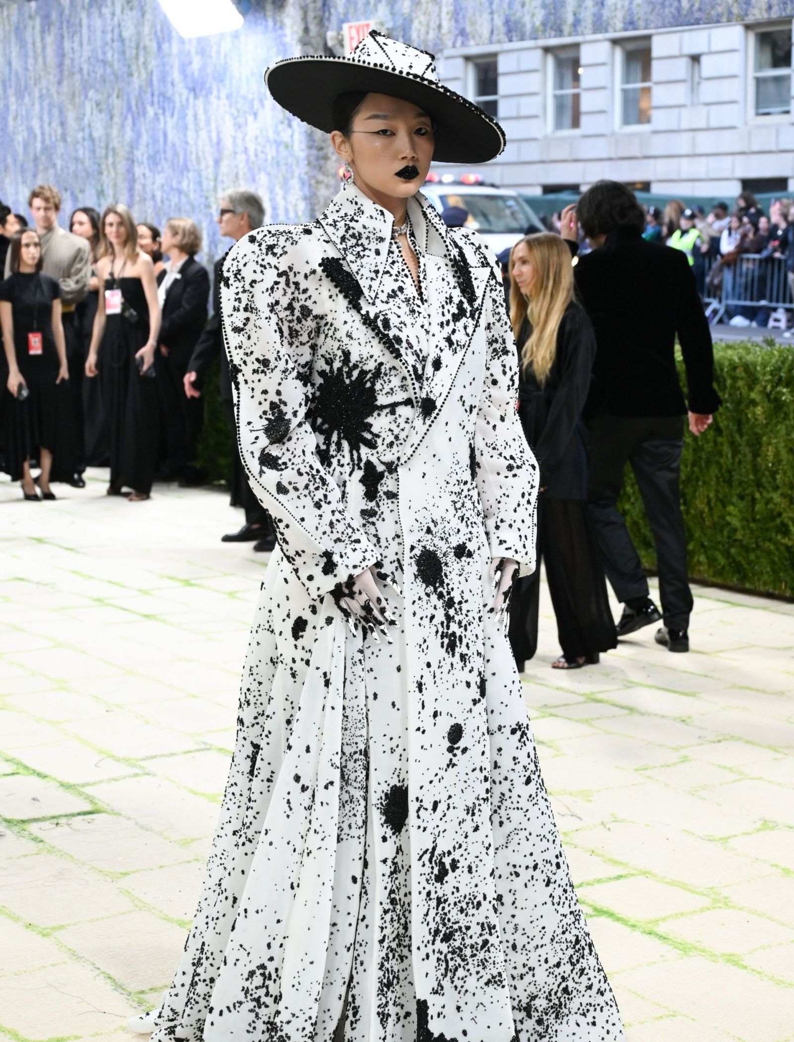 Model wearing a white gown with black paint-splatter pattern and a wide black hat at an outdoor fashion event