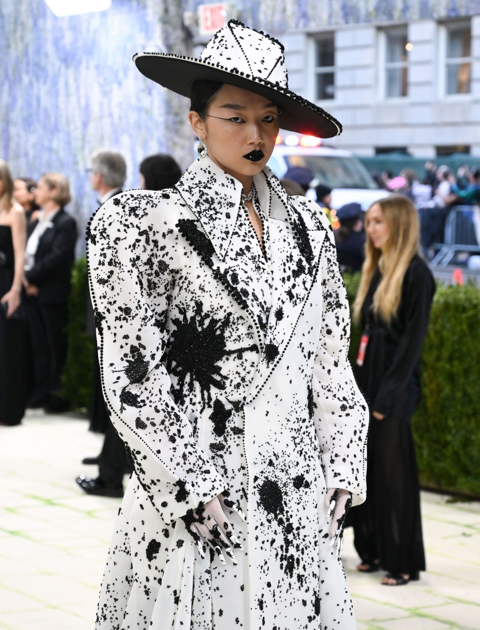 Model in a white speckled suit with a large black-and-white hat, bold black lipstick, and dramatic makeup at a fashion event.