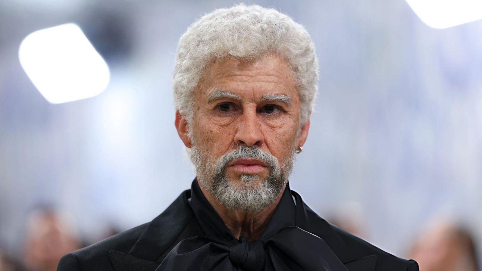 Older man with a white beard and mustache wearing a black suit and bow tie, looking straight ahead.
