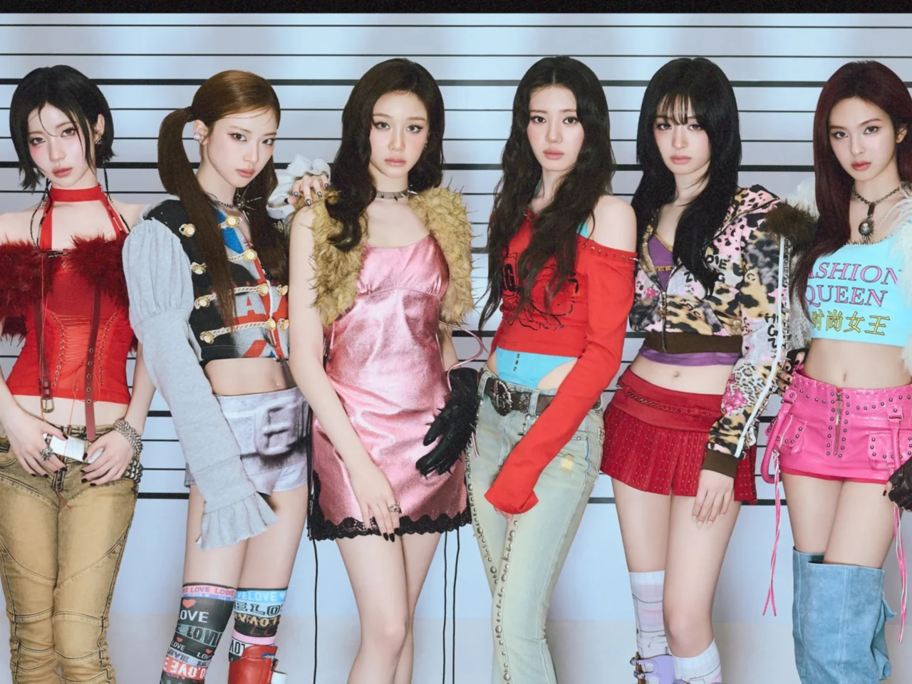 Seven female models standing in a line in colorful outfits against a striped height-chart backdrop, posing for a fashion photoshoot.