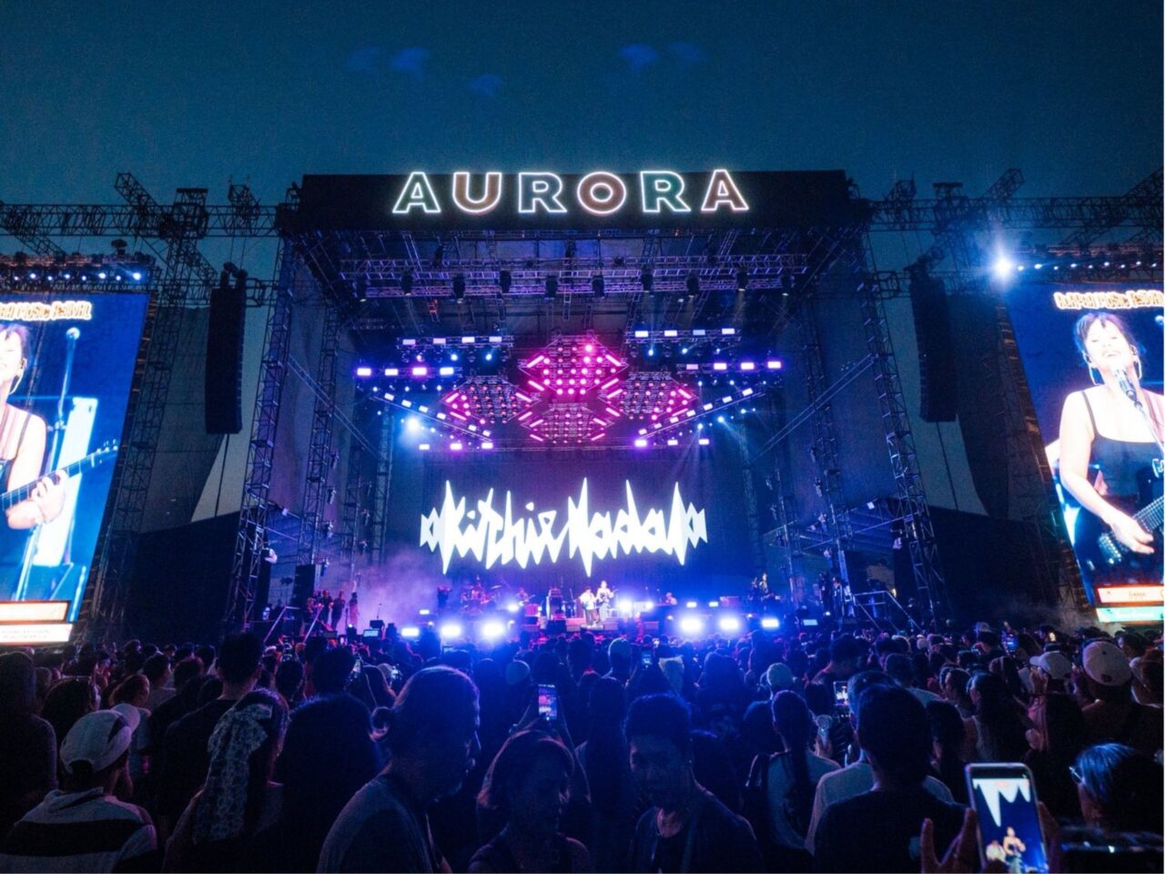 Wide nighttime concert scene with a huge stage, neon AURORA sign, and a cheering crowd against blue lights.