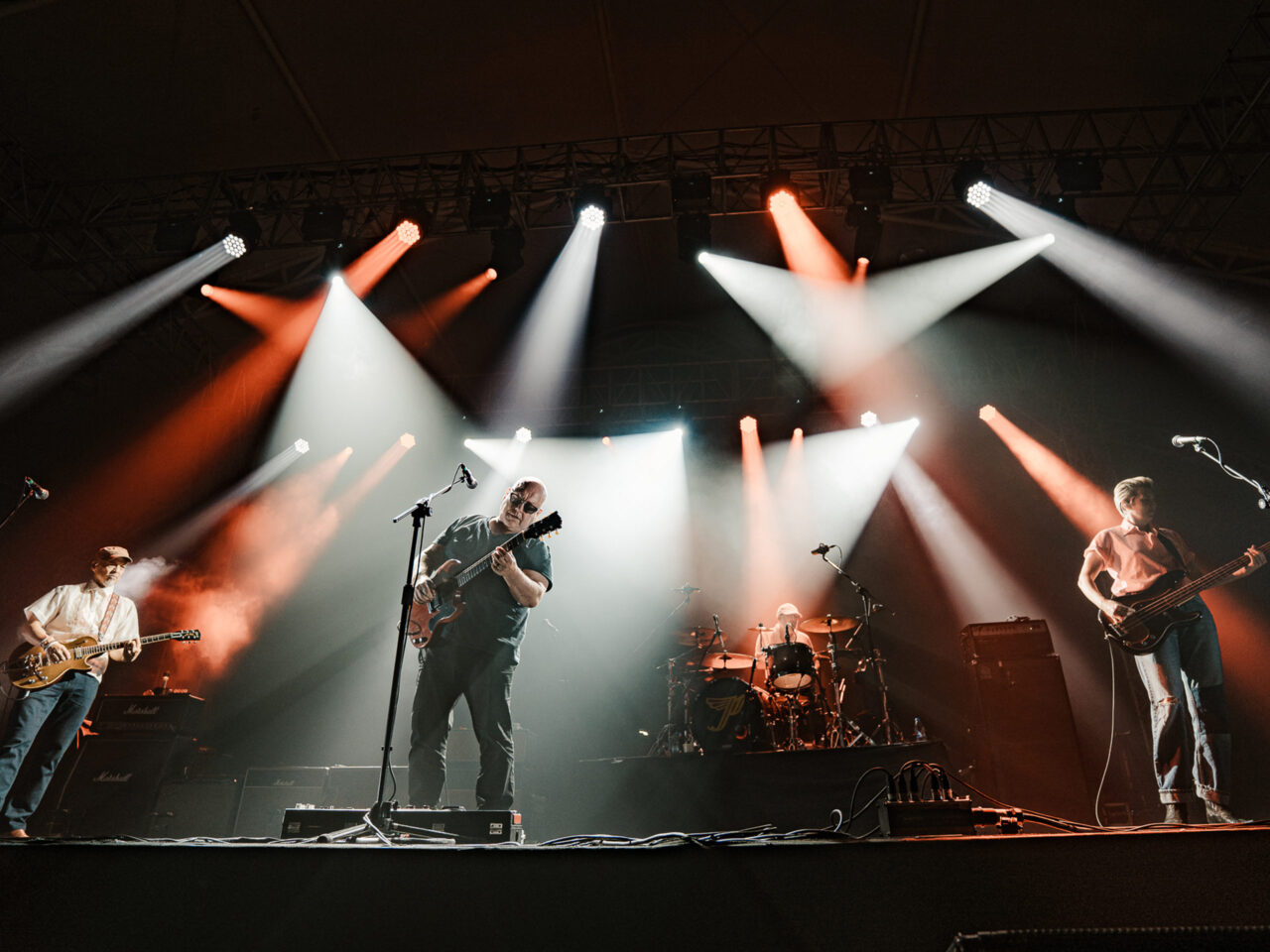 A rock band performs on a dark stage with red and white spotlights, guitarist center, bassist right, drummer in back.