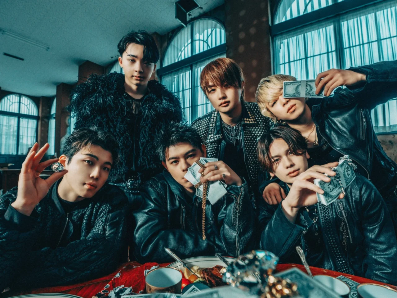 Six young men in leather jackets pose indoors, displaying cash in a moody, urban setting.