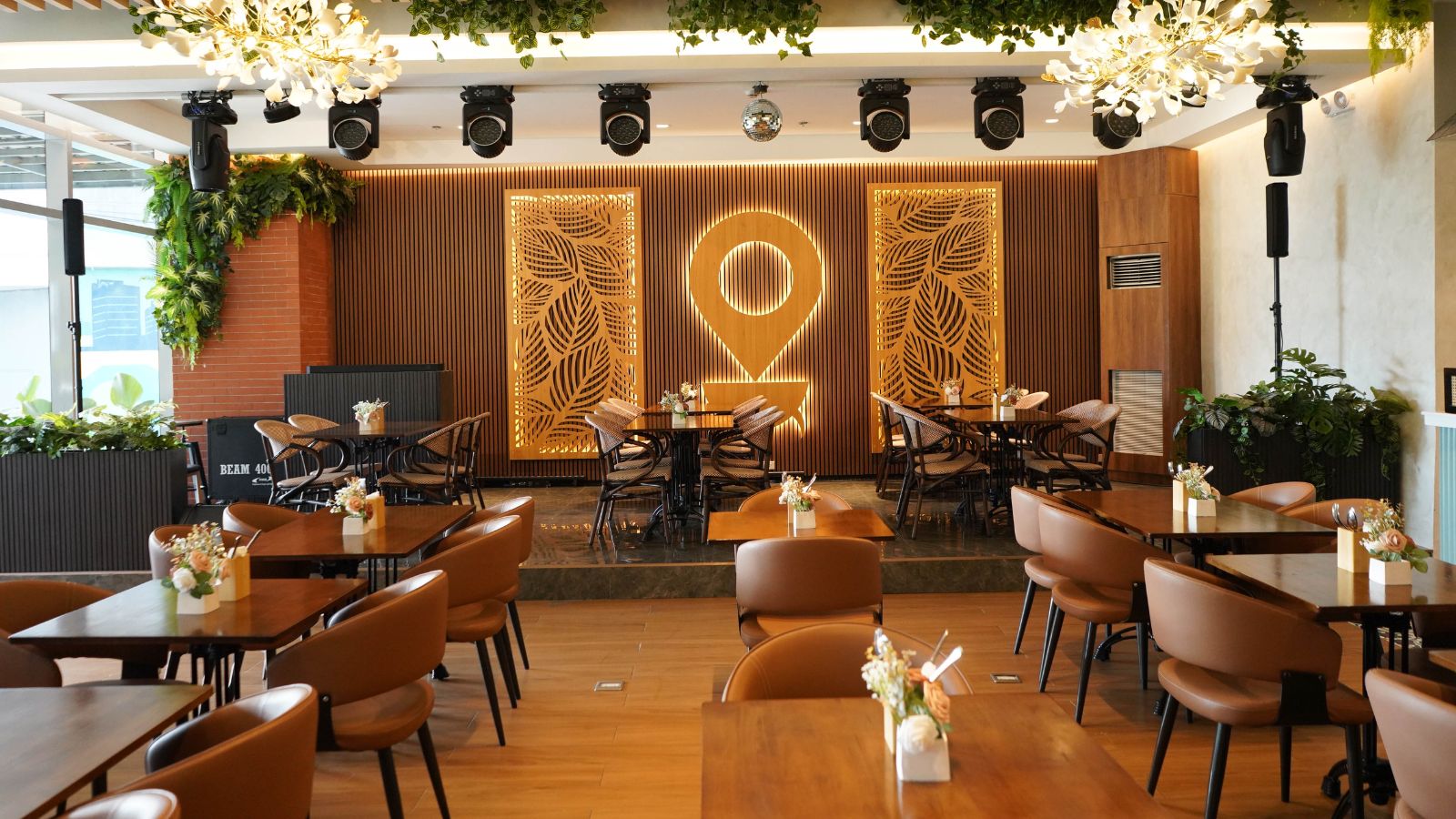 Elegant restaurant interior with warm wood panel backdrop, decorative gold panels, and a central pin-like design, greenery, and tables set with flowers.