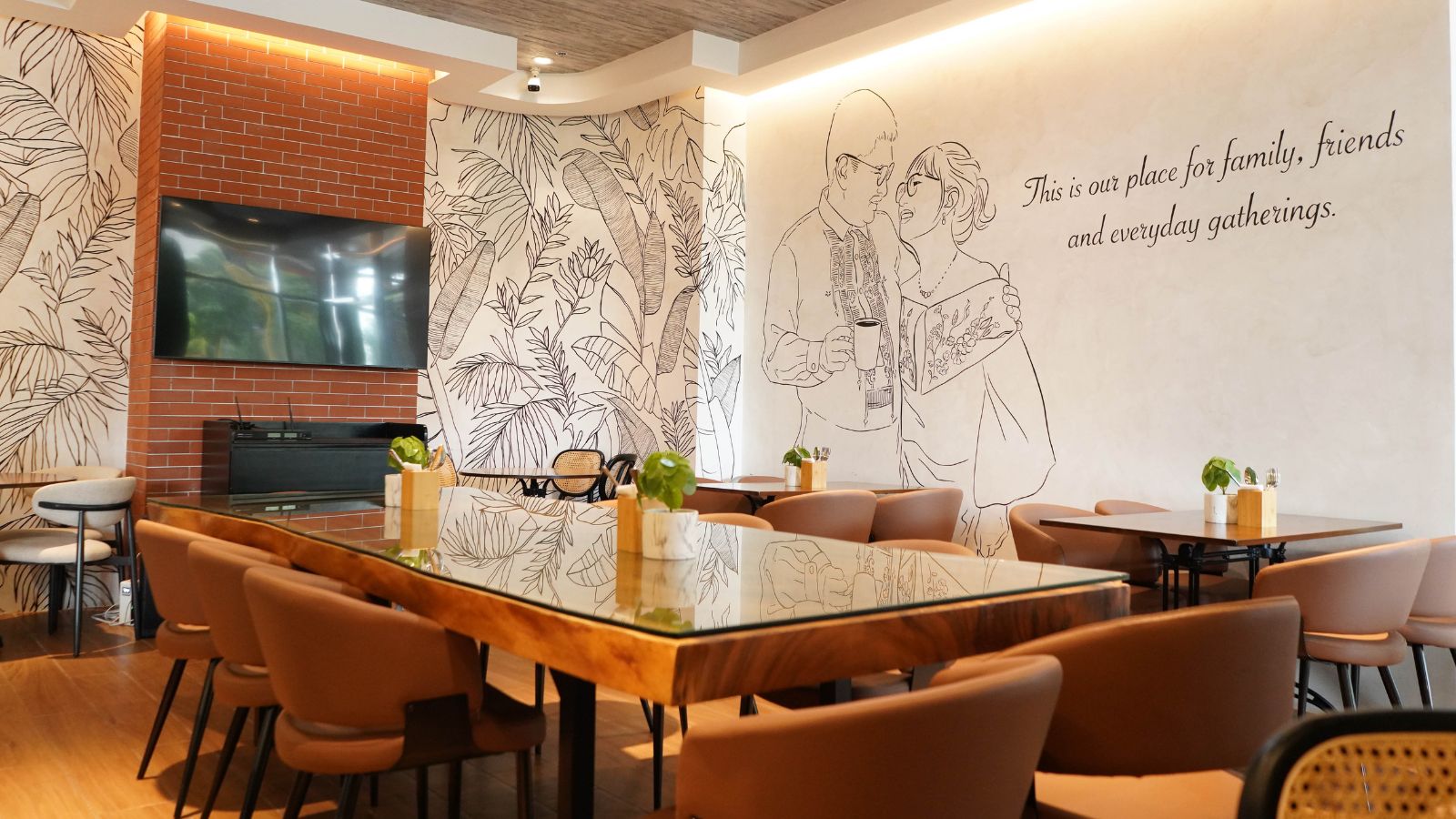 Modern cafe interior with a large mural, brick fireplace, and a long glass-top table surrounded by tan chairs.