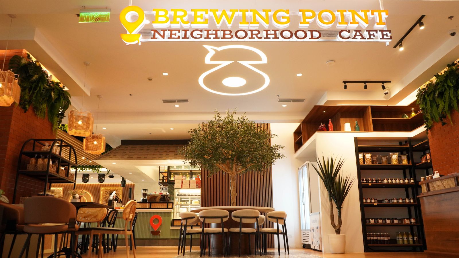 Interior of a cafe with a large illuminated sign reading 'BREWING POINT NEIGHBORHOOD CAFE', warm lighting, and a central tree surrounded by stools and tables.