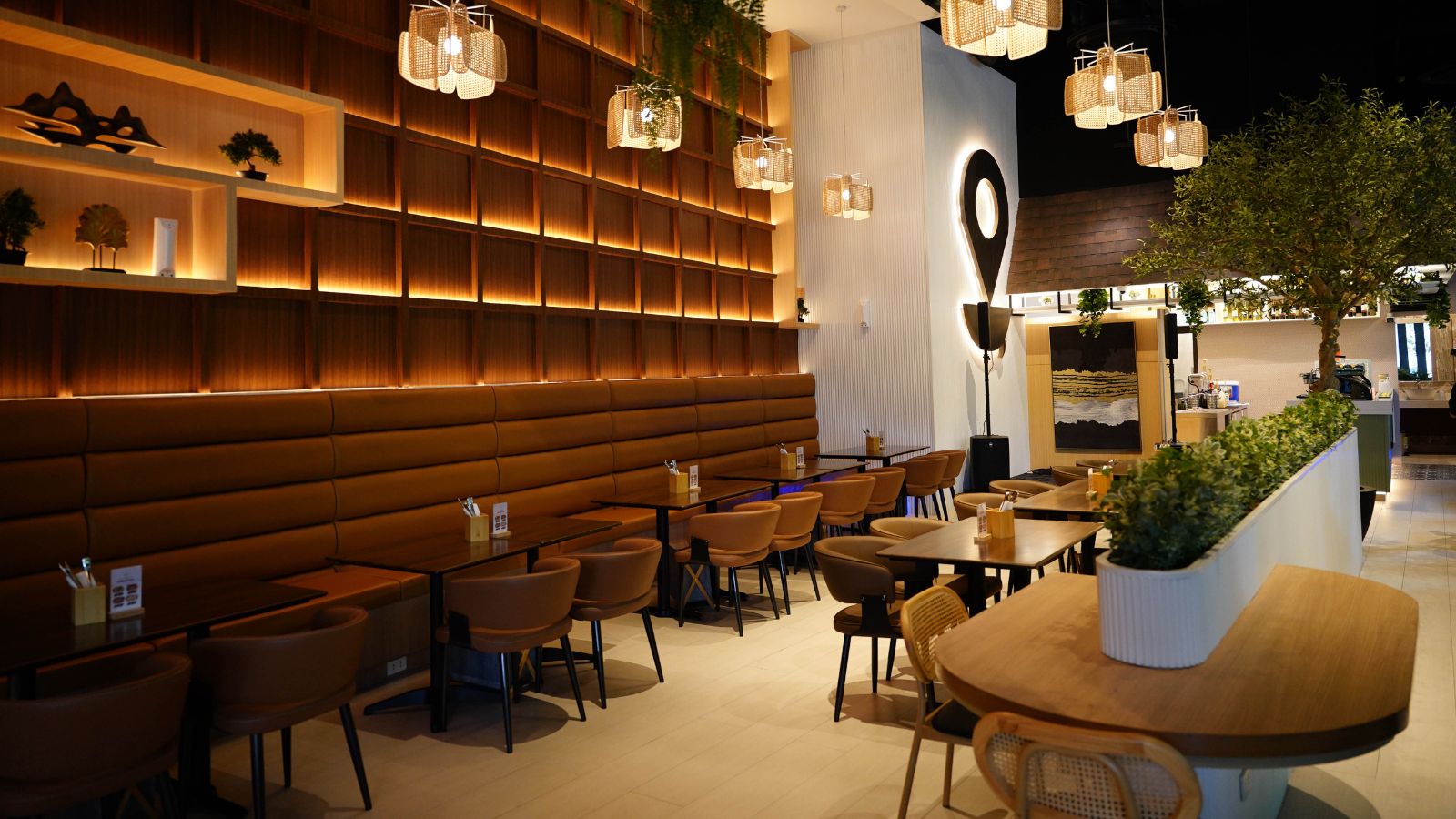 Modern cafe interior with a long brown leather banquette, wooden wall cubbies, round tables, wicker pendant lights, and potted plants.