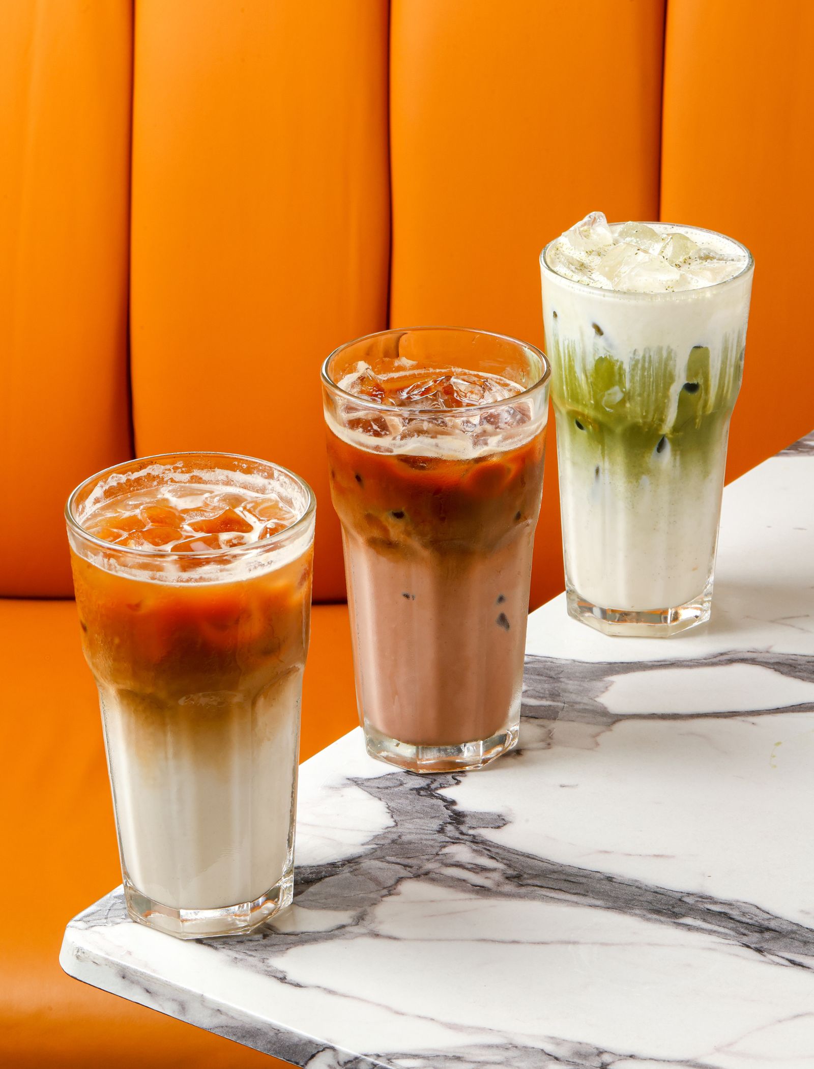 Four iced drinks in tall glasses on a marble counter with an orange background wall behind them.
