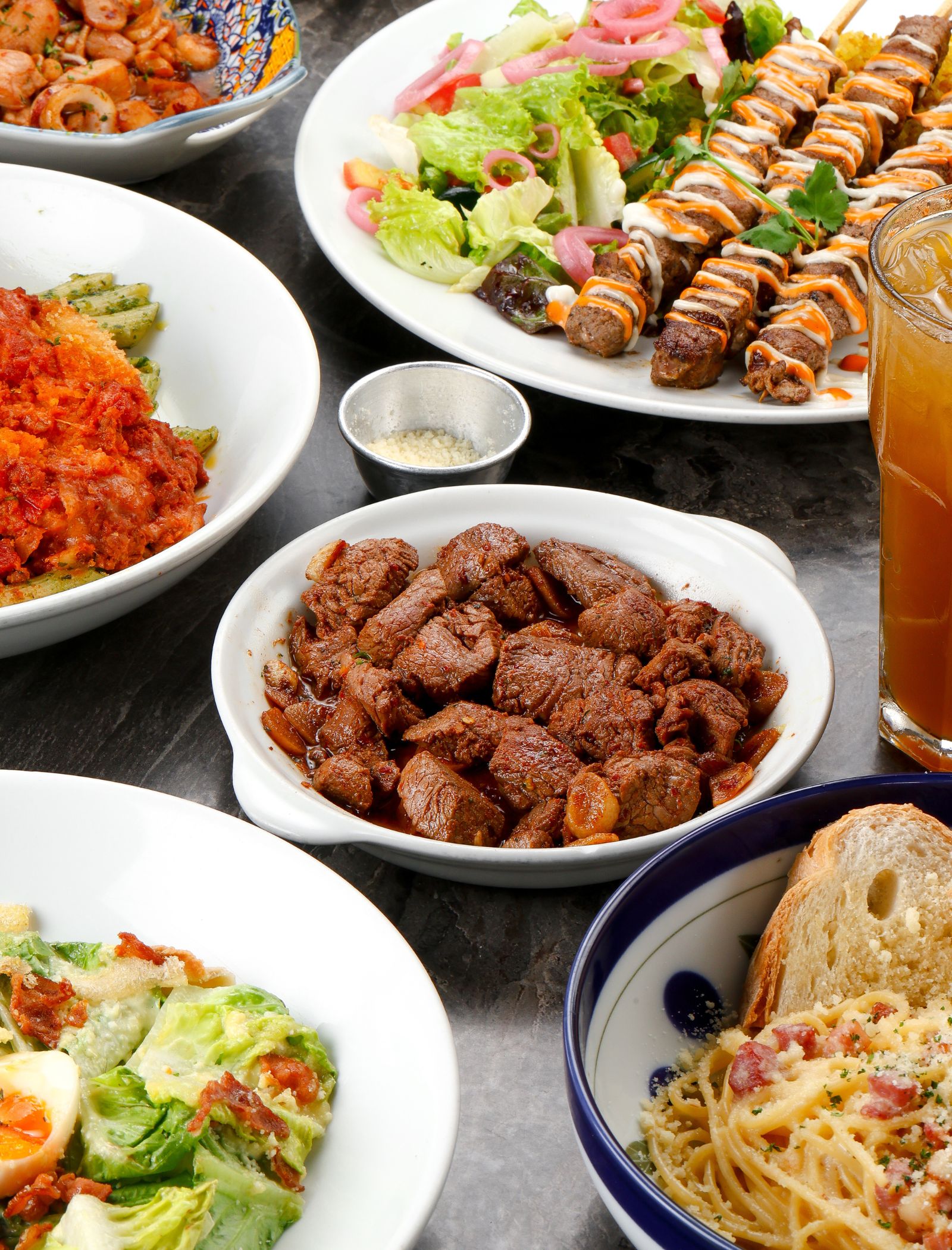 Bundle of dishes on a table: pasta with bacon, salad, grilled skewers, and beef chunks with a glass of iced drink nearby.
