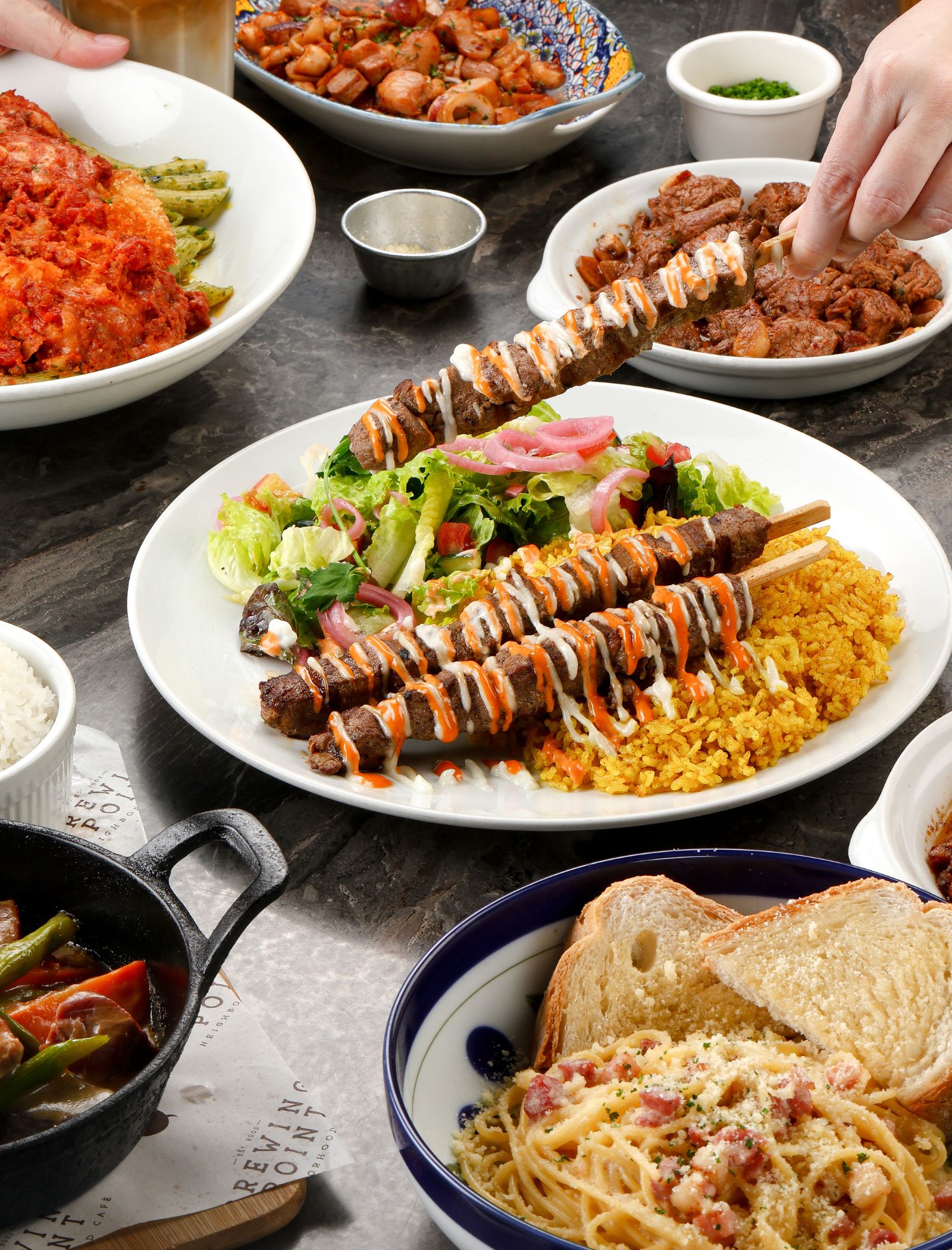 Assorted dishes on a table: skewered grilled meat with sauces over rice and greens, beside pasta and vegetables.