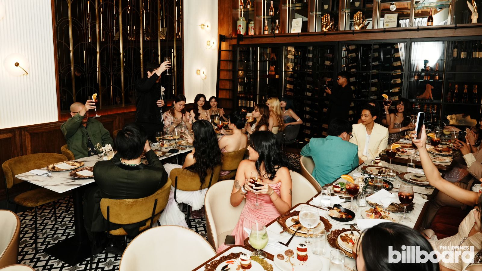 Group of people at a long dining table in an upscale wine-bar restaurant, toasting with cocktails and wine amid plates of dessert.