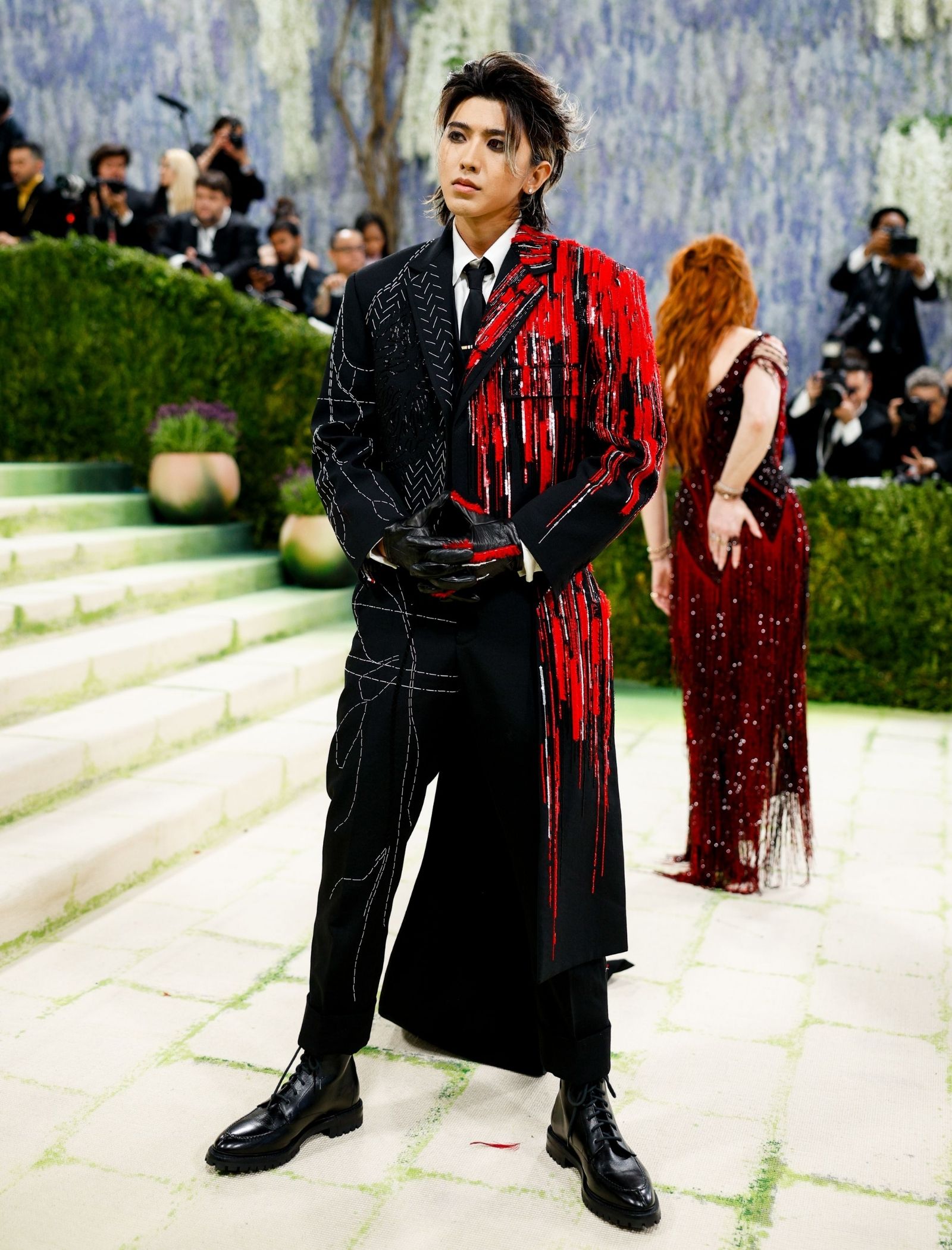 Model wearing a black-and-red split suit with fringed red fringe, standing on steps as photographers shoot nearby.