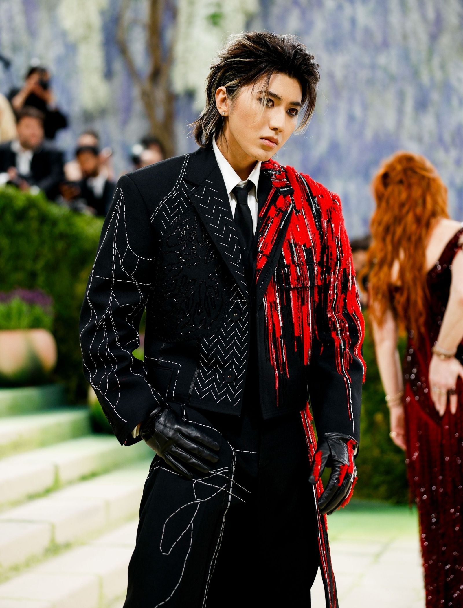 Fashion model wearing a black suit with white stitched patterns and a red textured jacket, walking on a runway outdoor event.