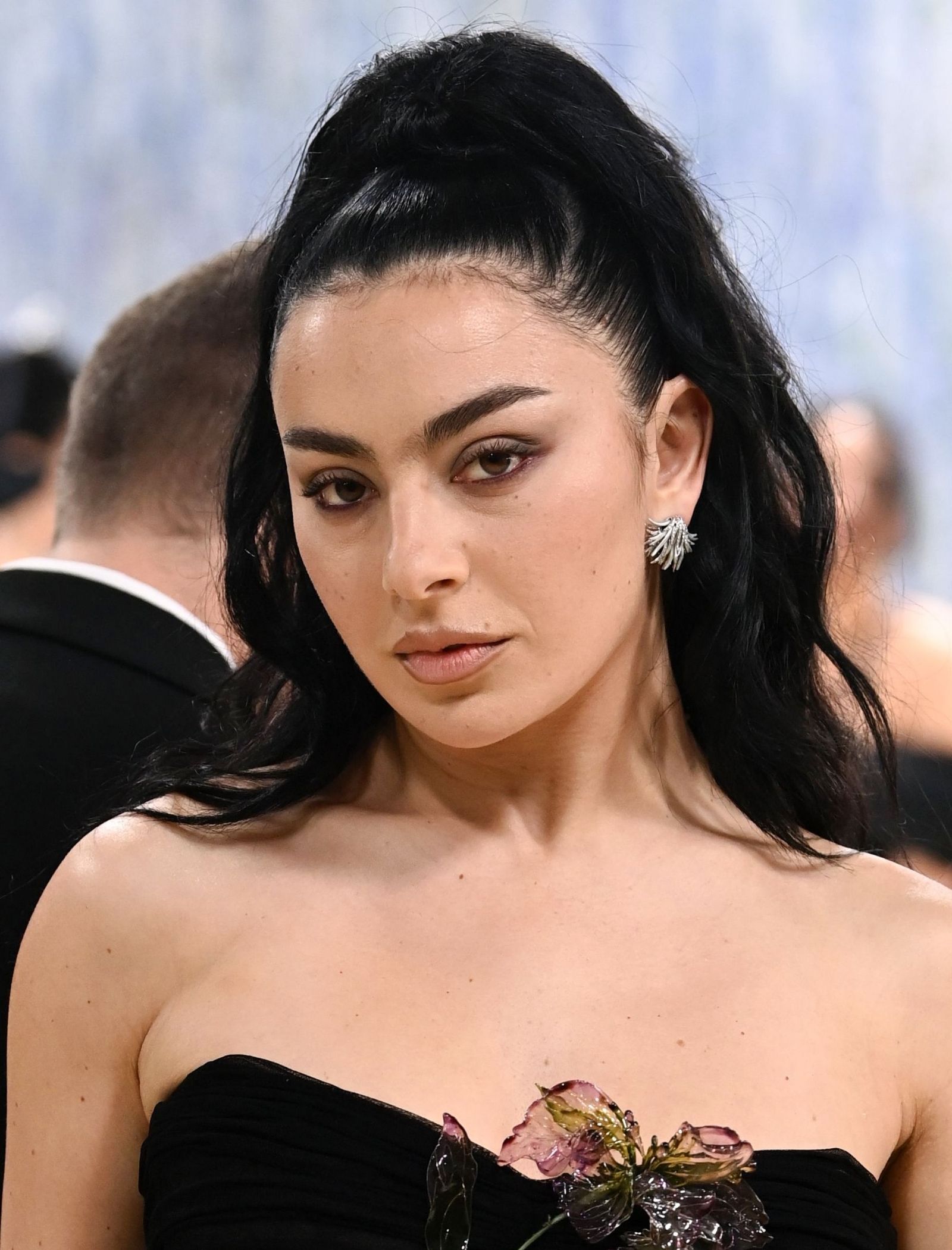Close-up of a woman with dark hair styled in a high ponytail, wearing a black strapless dress and a decorative floral corsage on the bodice with a silver leaf earring visible.
