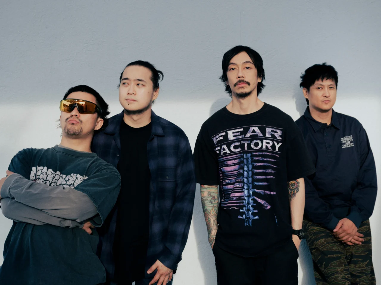 Four men pose against a plain gray wall in casual streetwear and relaxed expressions.