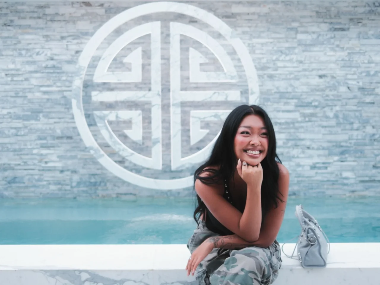 Smiling woman with long black hair crouches beside a pool, with a blue-gray stone wall and large circular logo behind her.