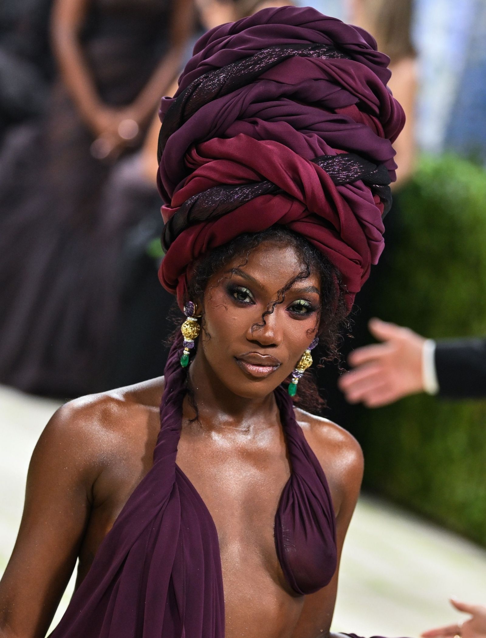 Portrait of a Black woman wearing a large burgundy headwrap and matching halter dress, gold earrings, outdoors.