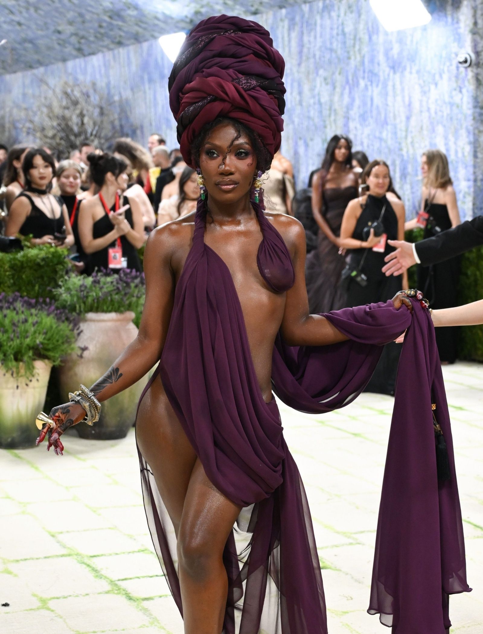 Model in a deep purple halter dress and oversized magenta headwrap posing on a fashion show runway with a crowd in the background.