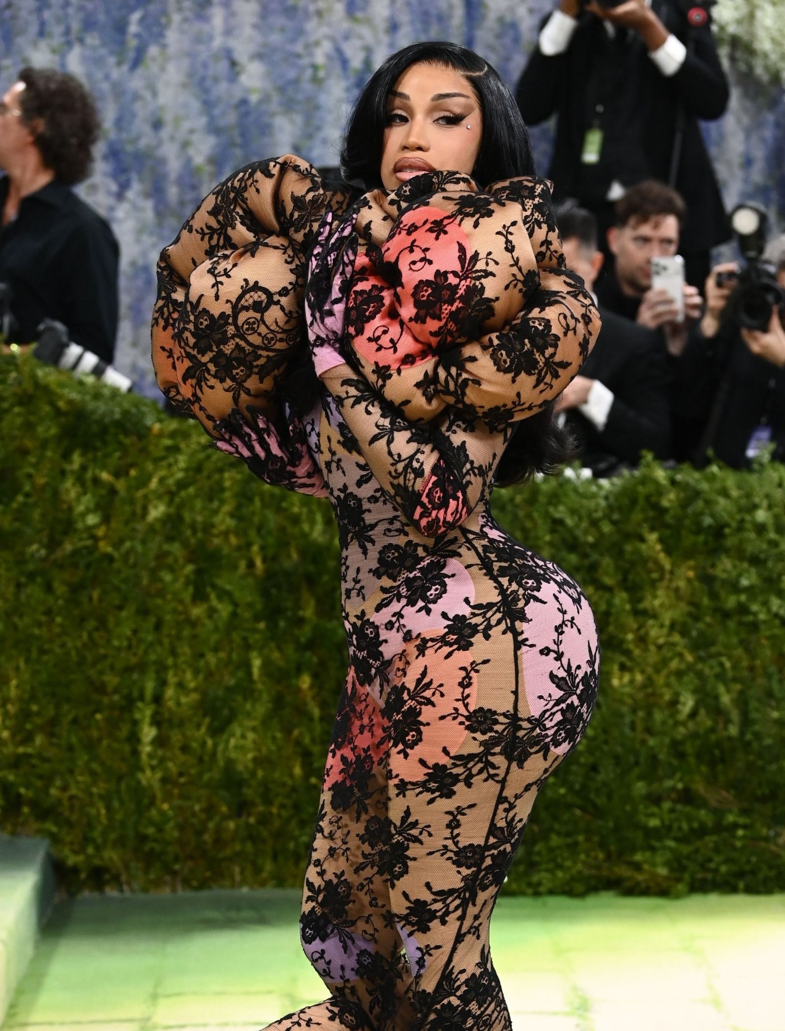 Person wearing a sheer black lace bodysuit with pink underlayer, posing on a green carpet as photographers snap photos in the background