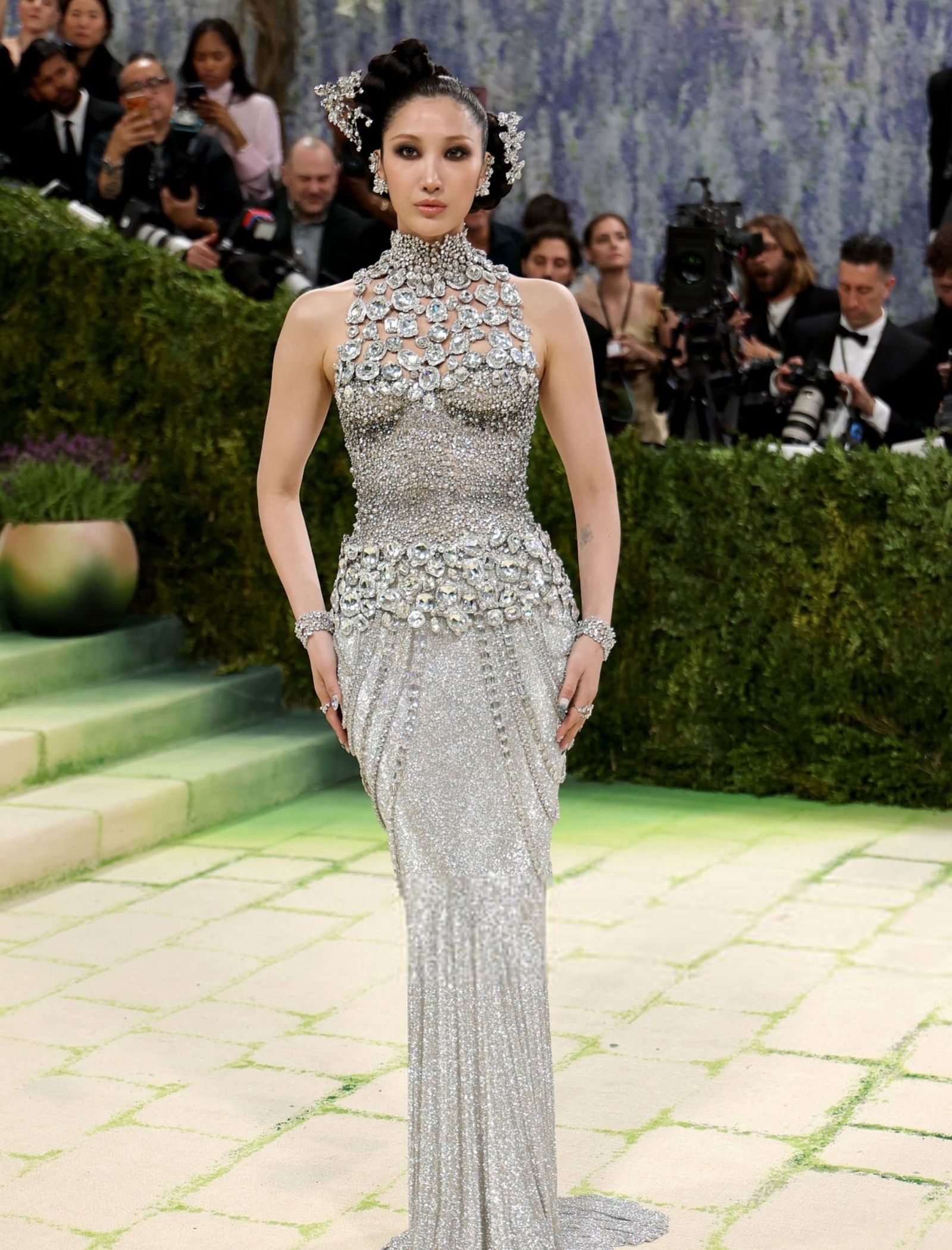 Glamorous woman in a sparkling silver jeweled gown with an ornate updo, posing on a green carpet as photographers capture her.