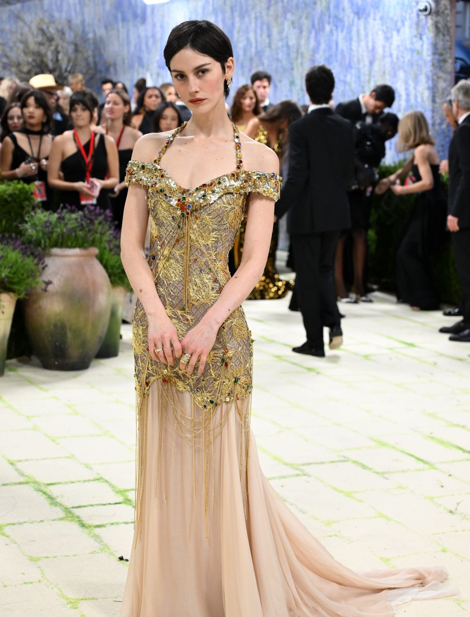 Female model in a gold beaded off-the-shoulder gown with a long, sheer train, standing on a fashion event runway with a crowd in the background.