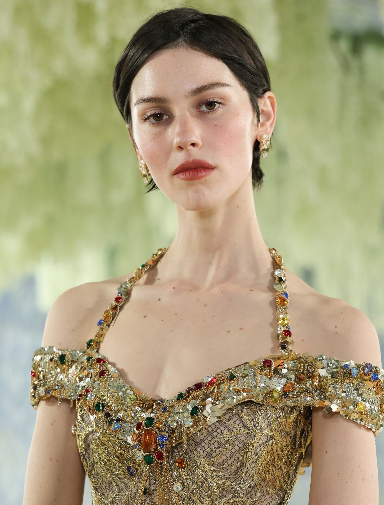 Portrait of a woman with short dark hair wearing a gold bejeweled dress and matching earrings against a blurred green background.
