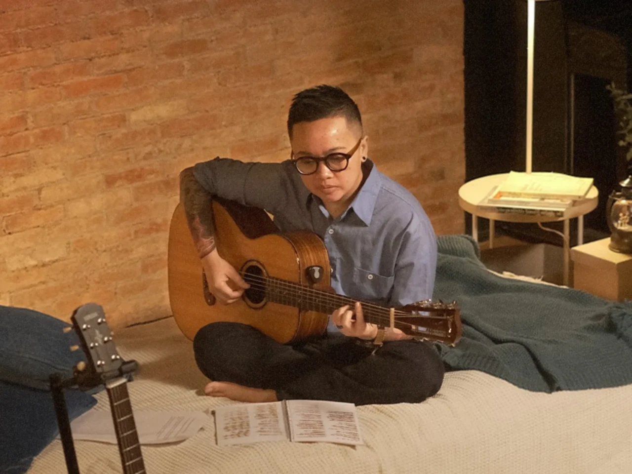 Person with glasses sits cross‑legged on a bed, playing an acoustic guitar in a brick-walled room.