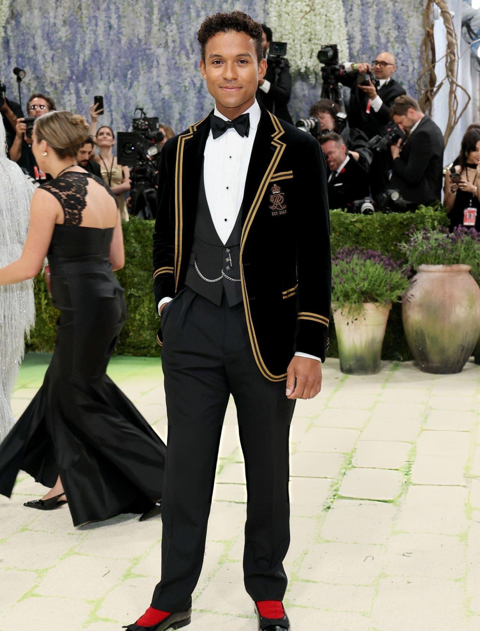 Young man in a black tuxedo with gold trim and bow tie posing on a photographer-filled red carpet backdrop.