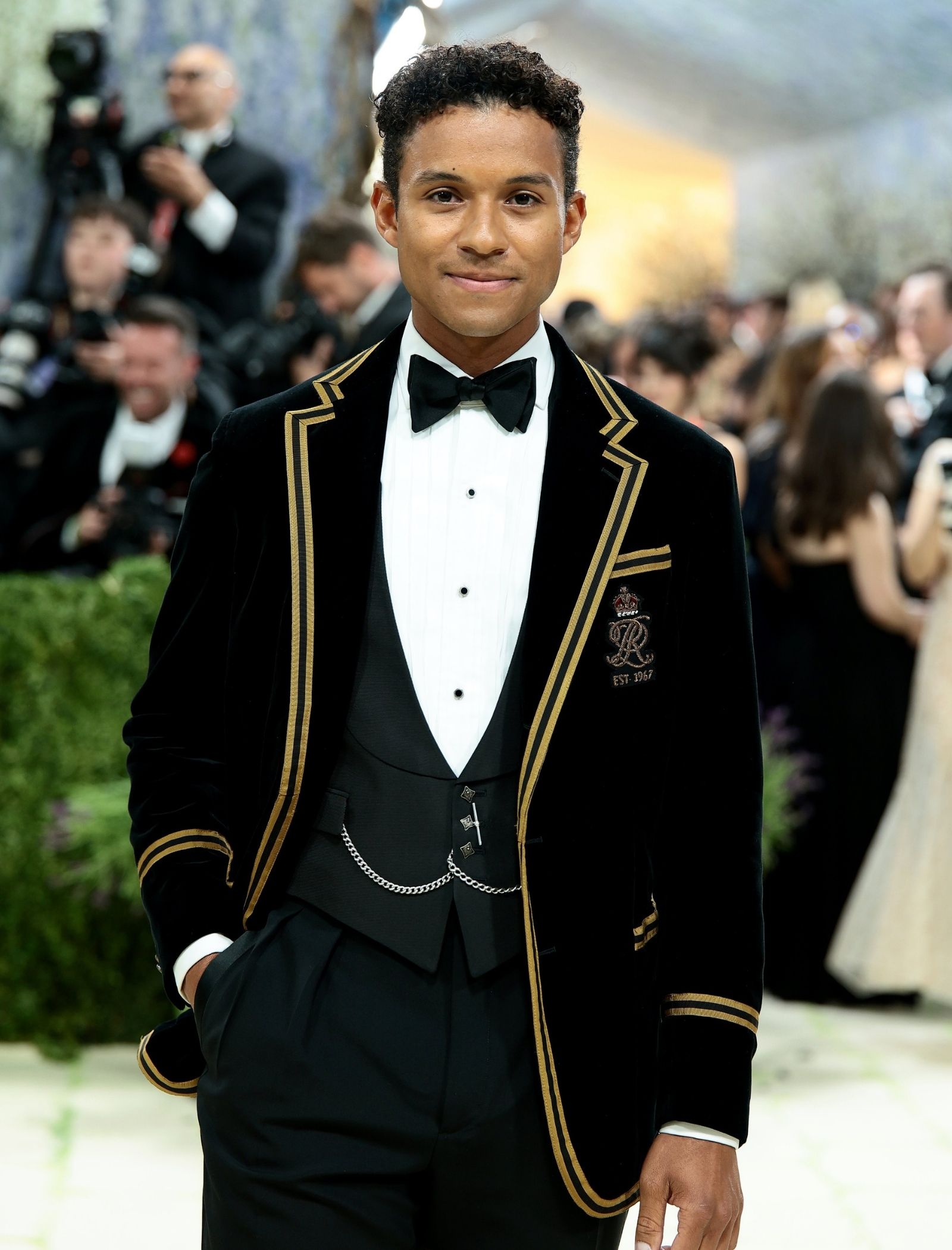 Young man in a black velvet tuxedo with gold piping and bow tie posing for photographers at a formal event.