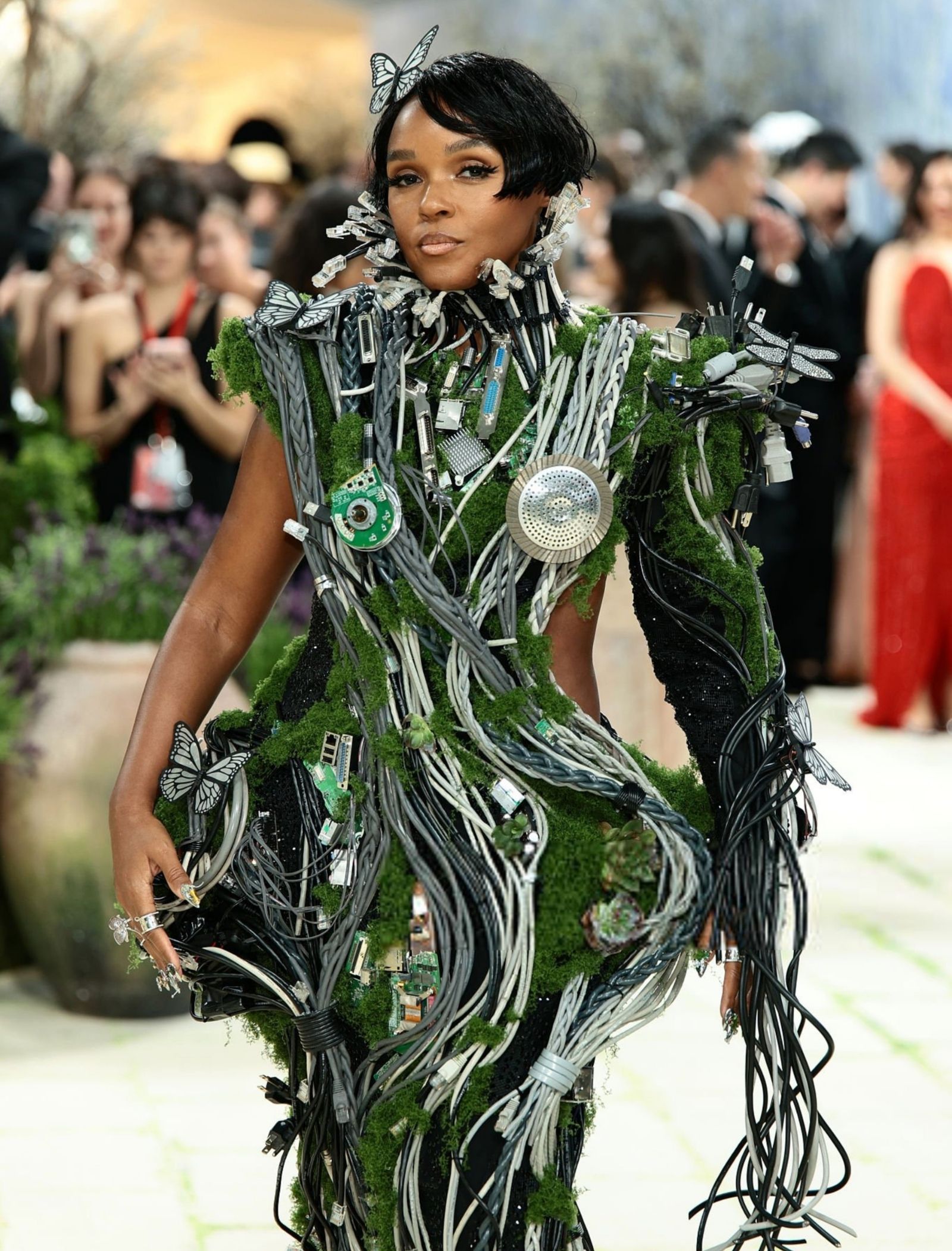 Model in an avant-garde dress made of cables and moss with butterfly accents at a red carpet event.