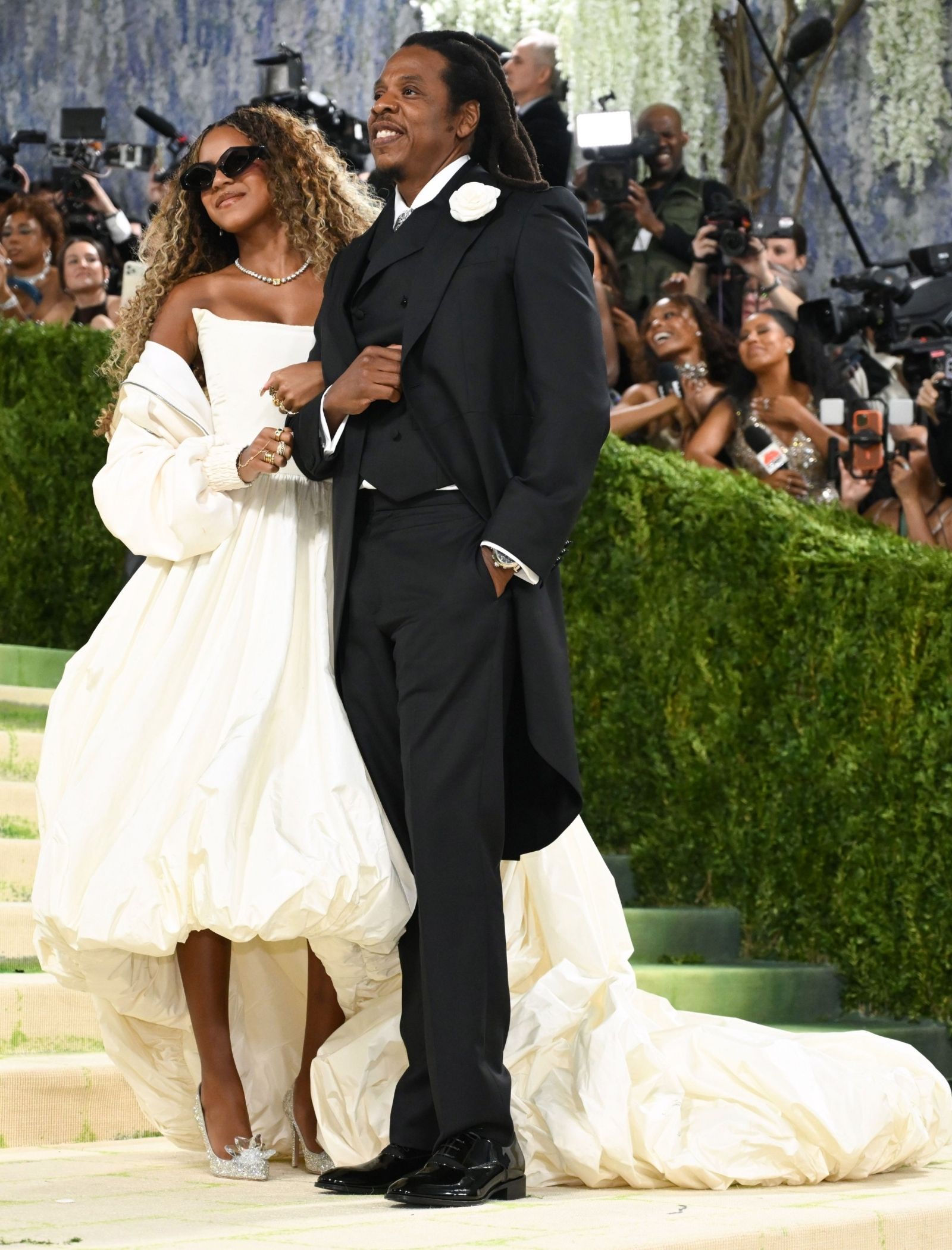 Smiling couple on a red carpet: woman in a white off-shoulder gown with a voluminous train and sunglasses, man in a black tuxedo with a white boutonniere, surrounded by photographers.