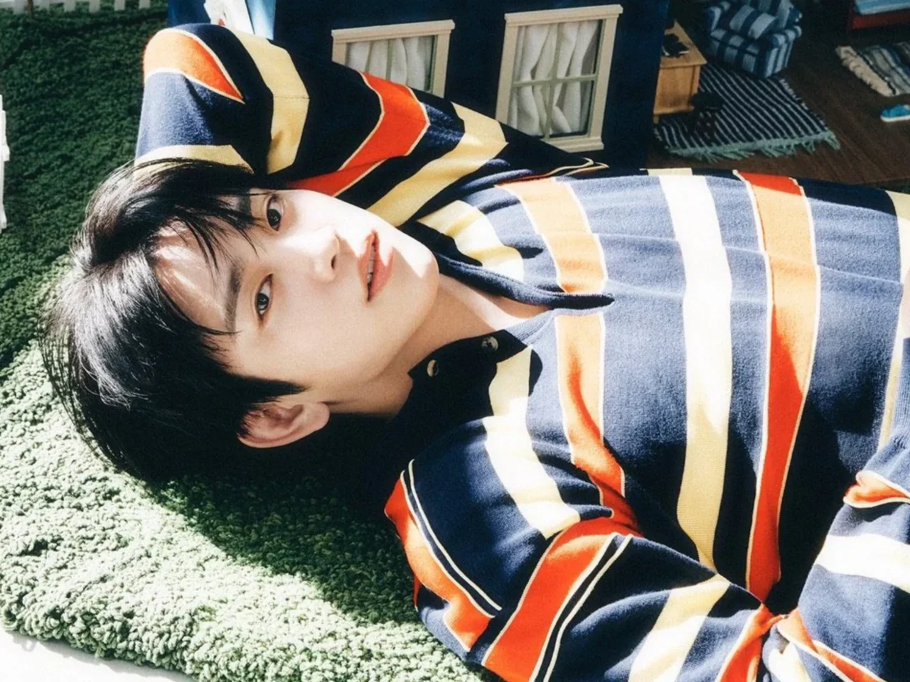 Young man with dark hair lying on a green shag rug, wearing a navy, orange, cream striped polo shirt.