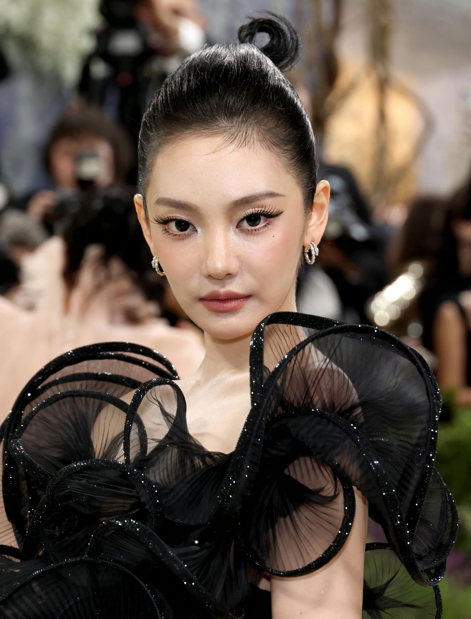 Close-up of a woman with a high black bun, bold makeup, and a black sheer ruffled dress at a fashion event.