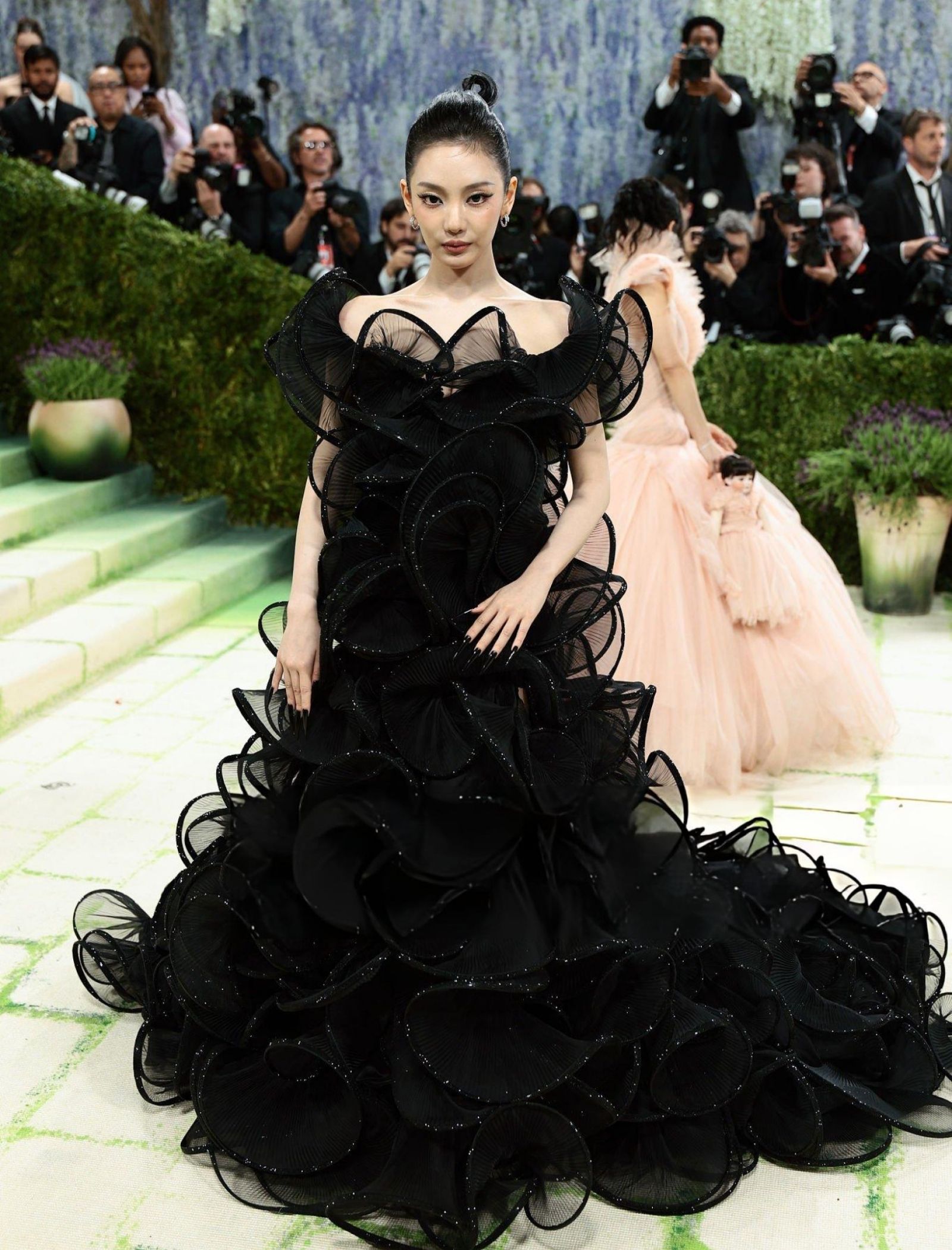 Asian model wearing a dramatic black ruffled gown on a green carpet as paparazzi flash behind her