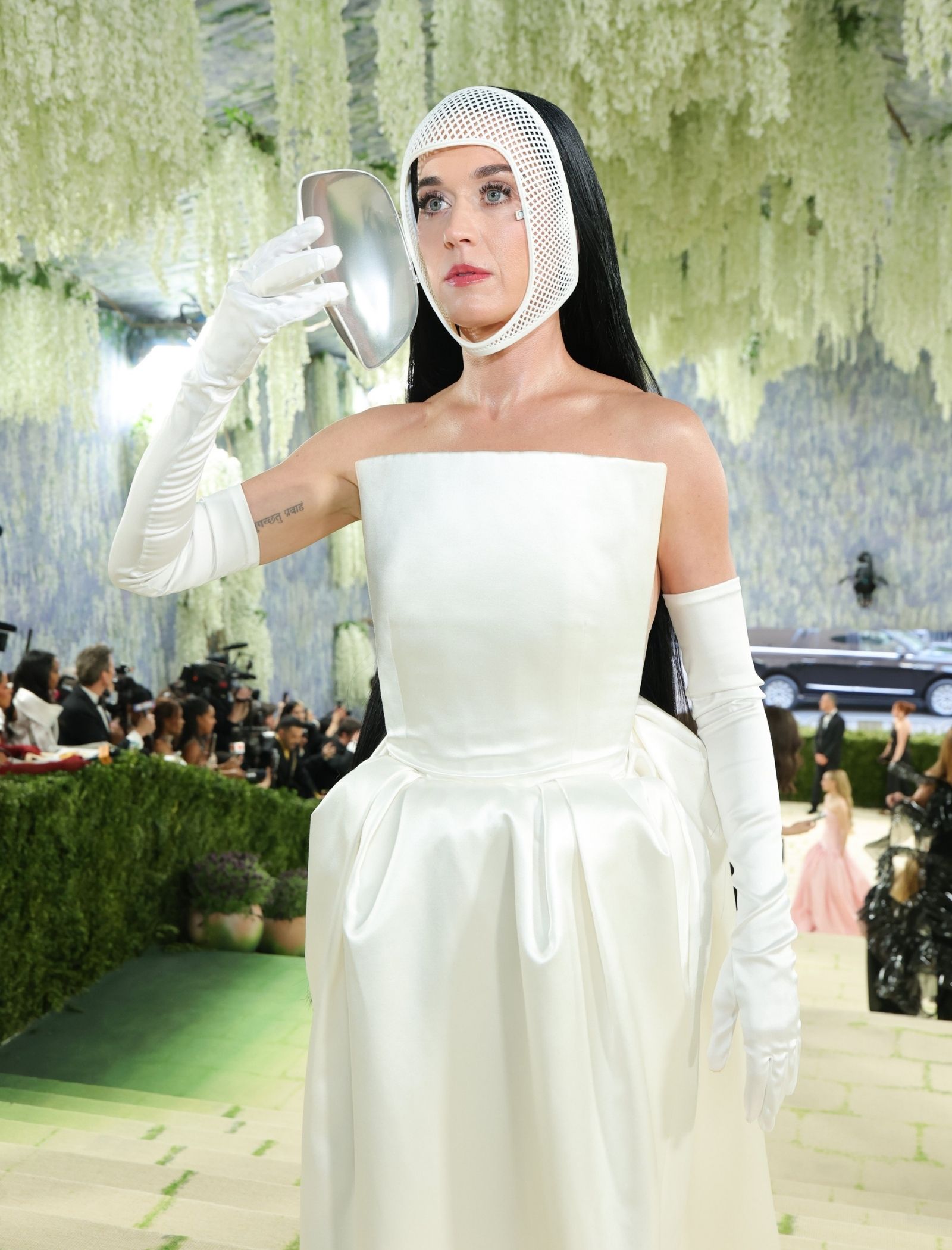 Model in a white satin gown with a perforated mesh hood, holding a round handheld mirror at a fashion event with greenery backdrop.