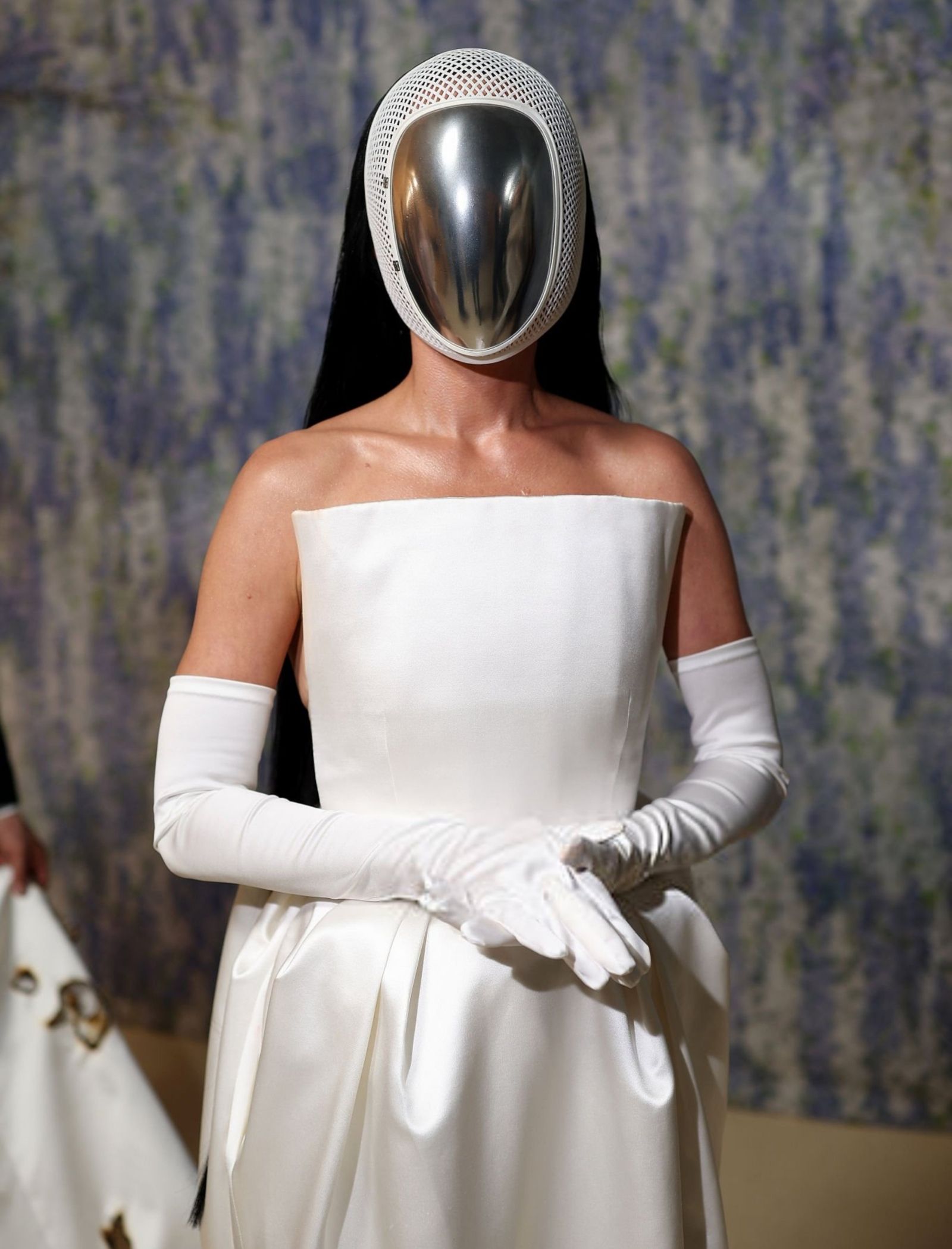 Model wearing a white off-shoulder gown with a shiny metal mask and white mesh hood on a runway fashion show stage.
