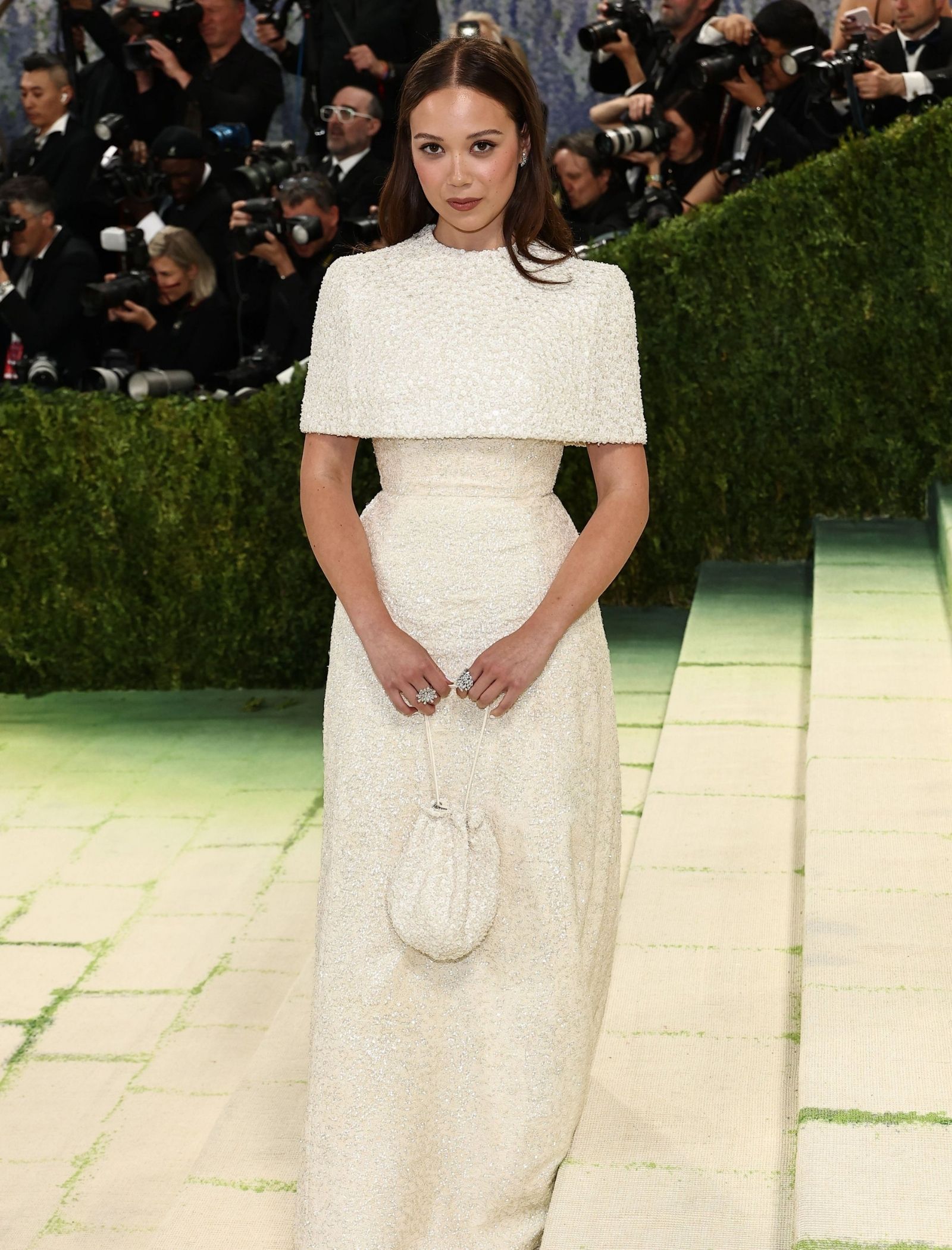 Woman in a long white beaded dress posing on a green carpet, surrounded by photographers at a red-carpet event.