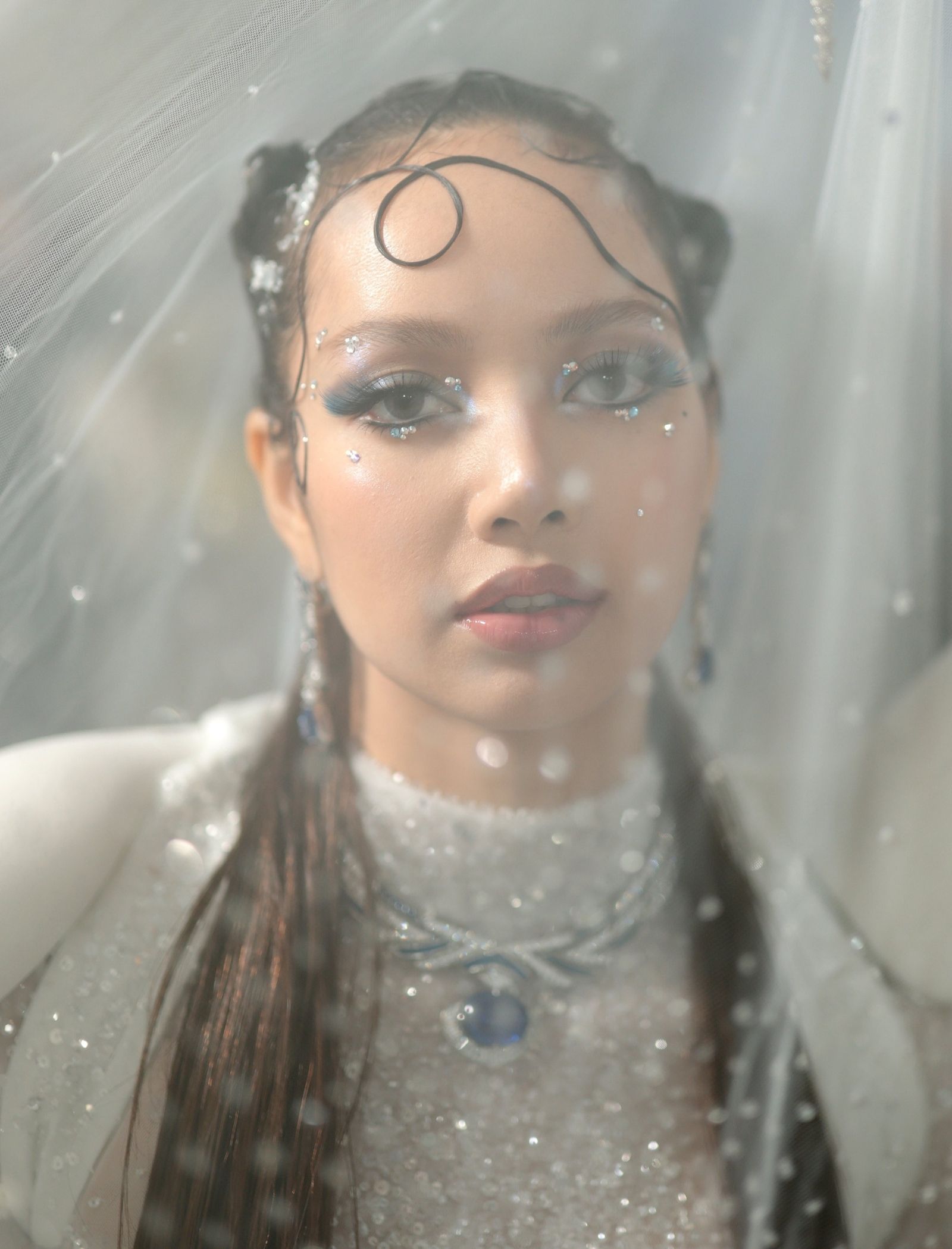 Close-up of a woman's face with blue glitter eye makeup and rhinestones under a sheer veil.