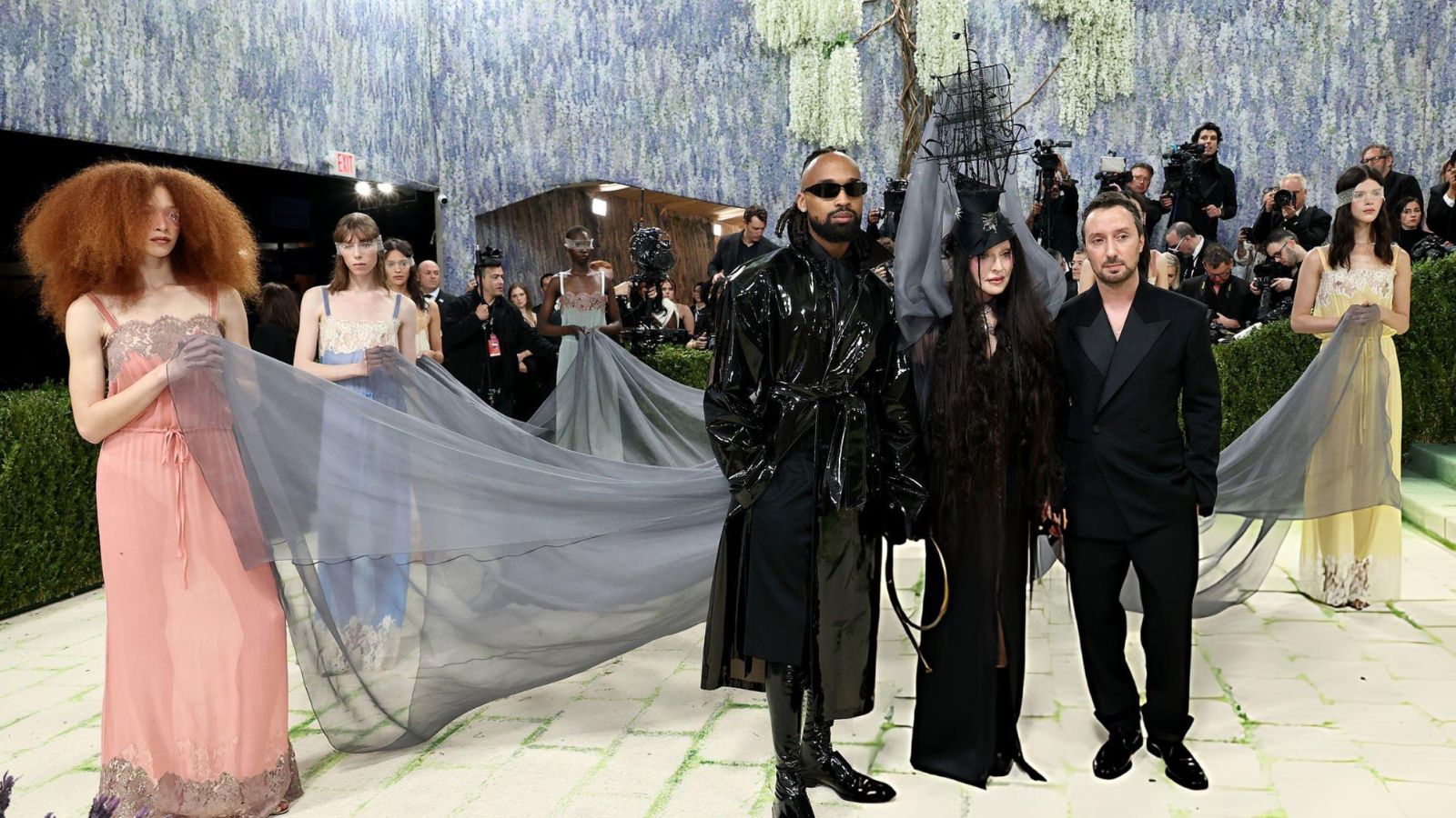Group of models and designers posing on a fashion gala red carpet, holding a long translucent gray train between them.