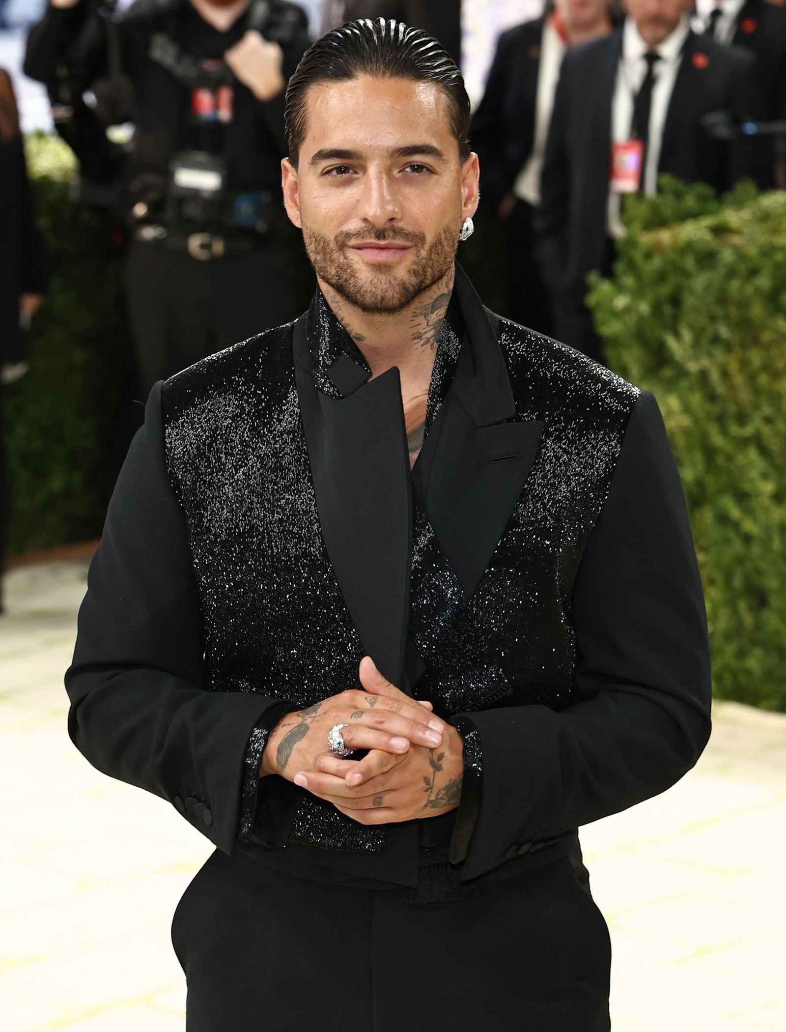 Man with slicked-back hair and stubble wearing a black glittery blazer, hands clasped in front at a formal event.
