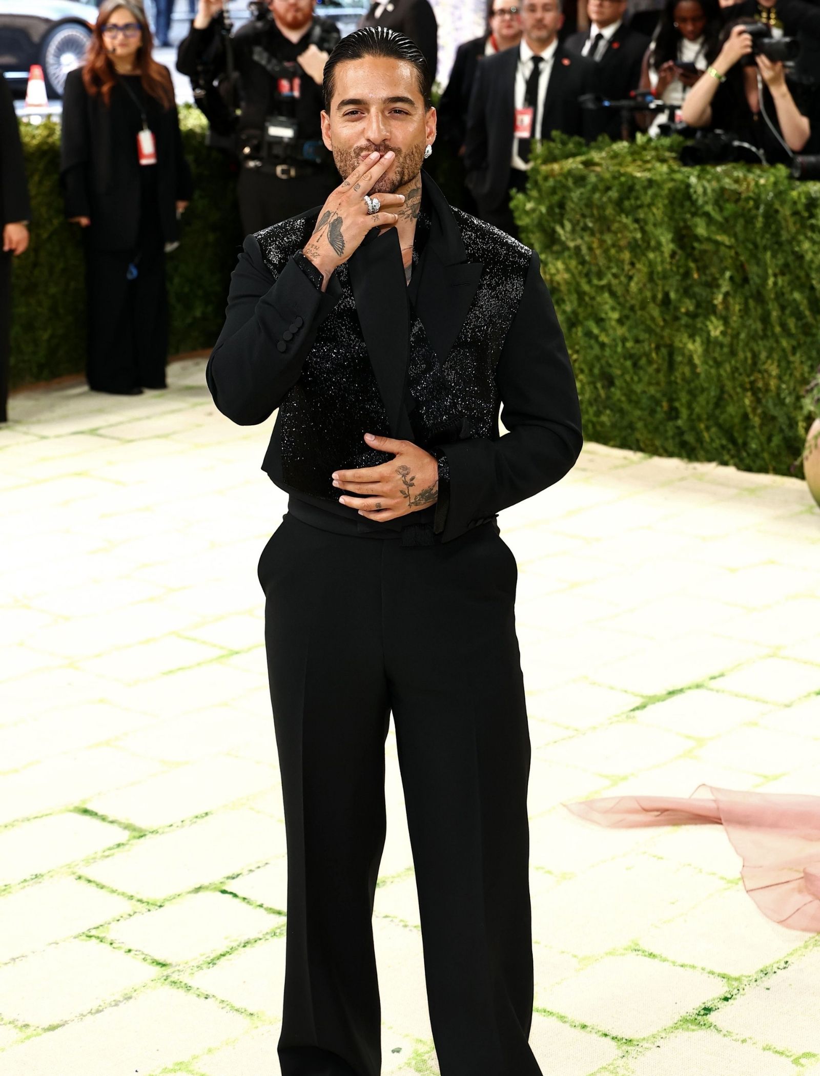 Man in a black sequined blazer posing for photographers, blowing a kiss with tattooed hands and rings visible.