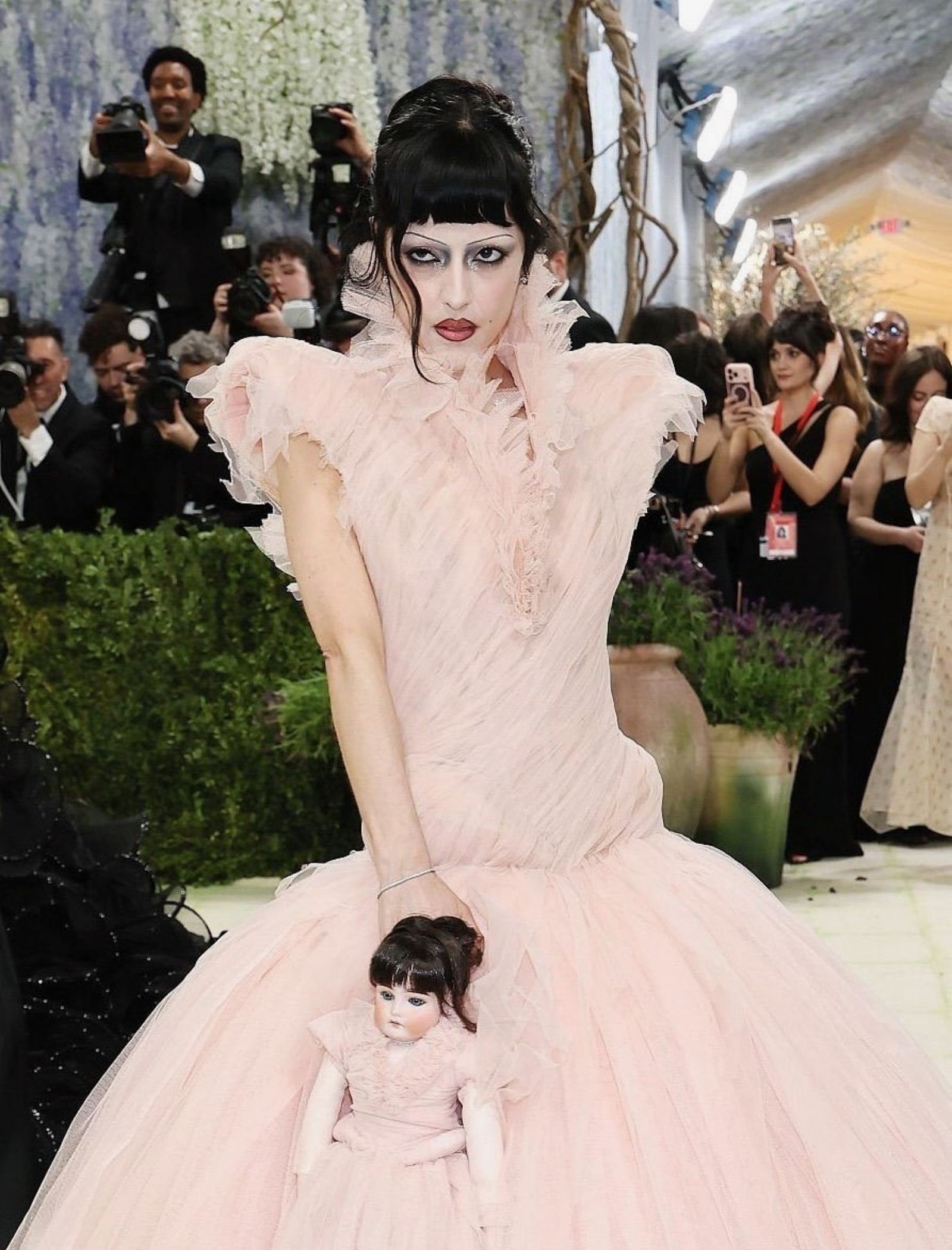 Model in a pale pink, voluminous gown posing for photographers; a small porcelain doll sits inside the dress folds near her waist area.